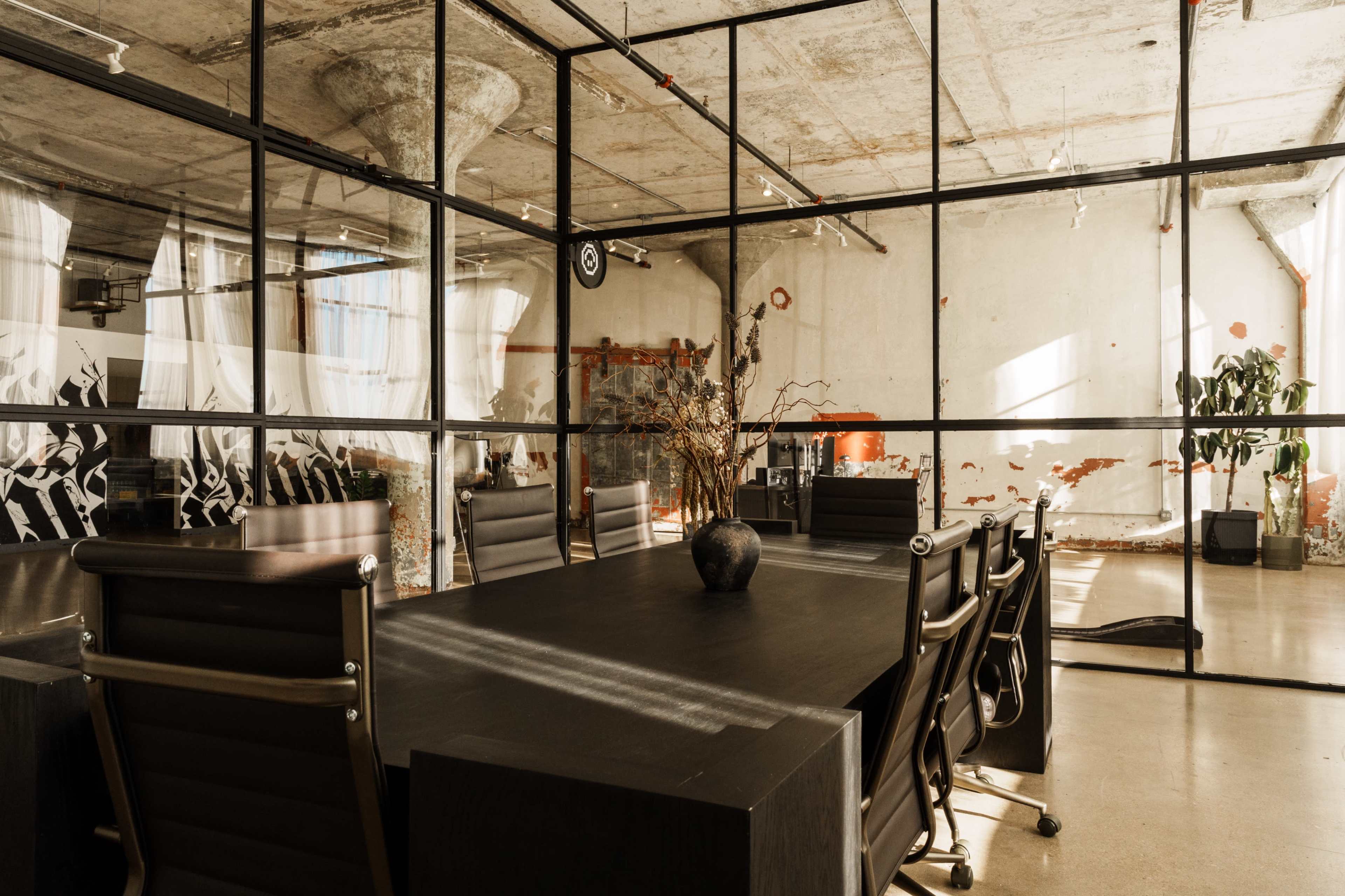 The image shows a modern conference room with a large black table surrounded by ergonomic chairs, enclosed by glass walls in a minimalist office space featuring exposed concrete and artistic decor.