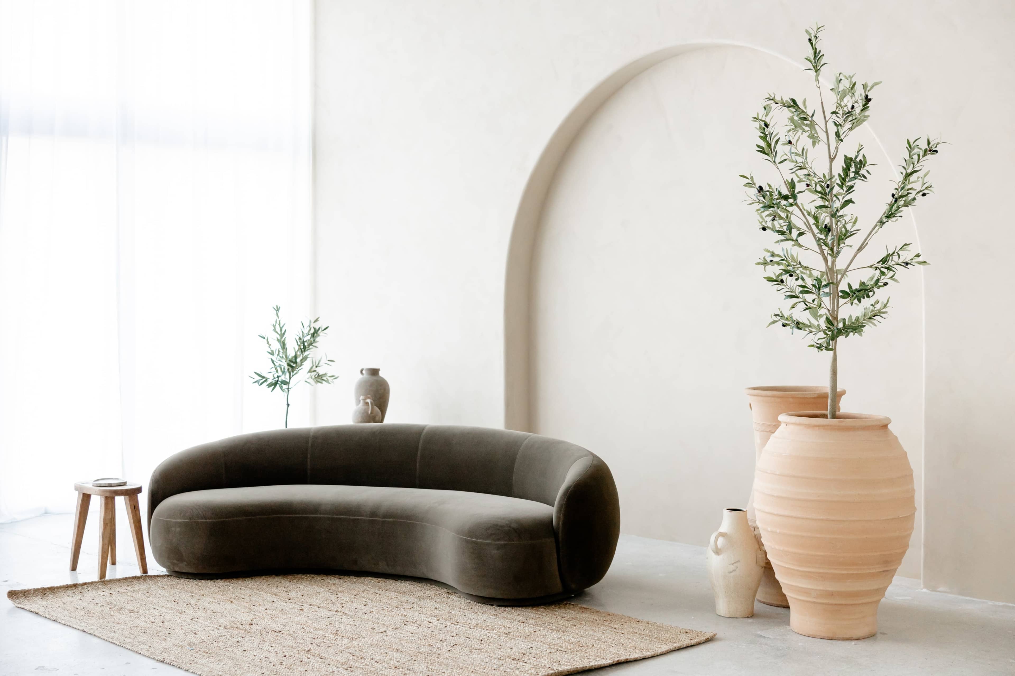 A curved sofa is positioned next to two large planters and a small side table in a minimalist interior space with a light-toned wall.