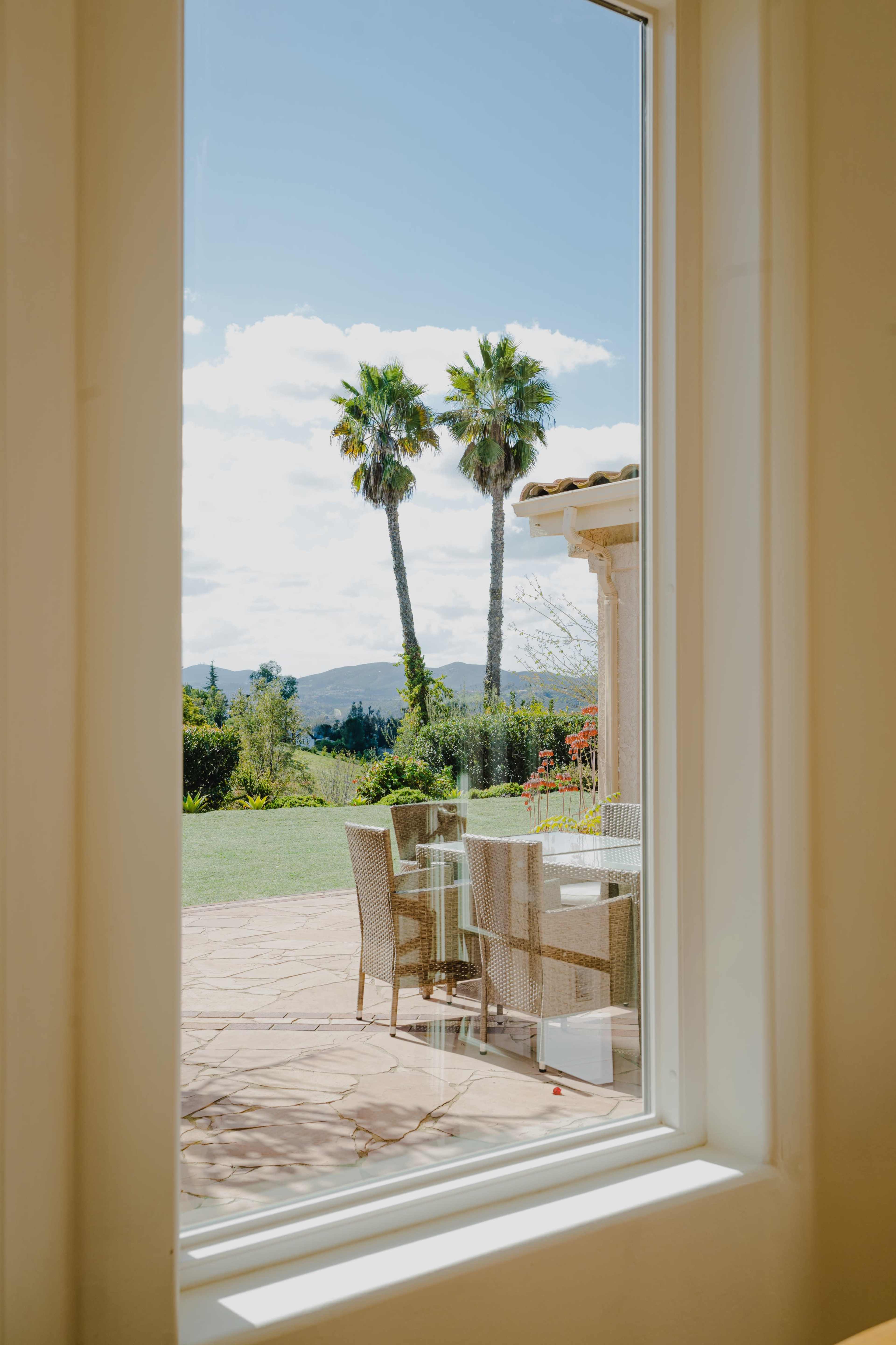 A clear view through a window shows a patio with wicker furniture and two palm trees against a blue sky.