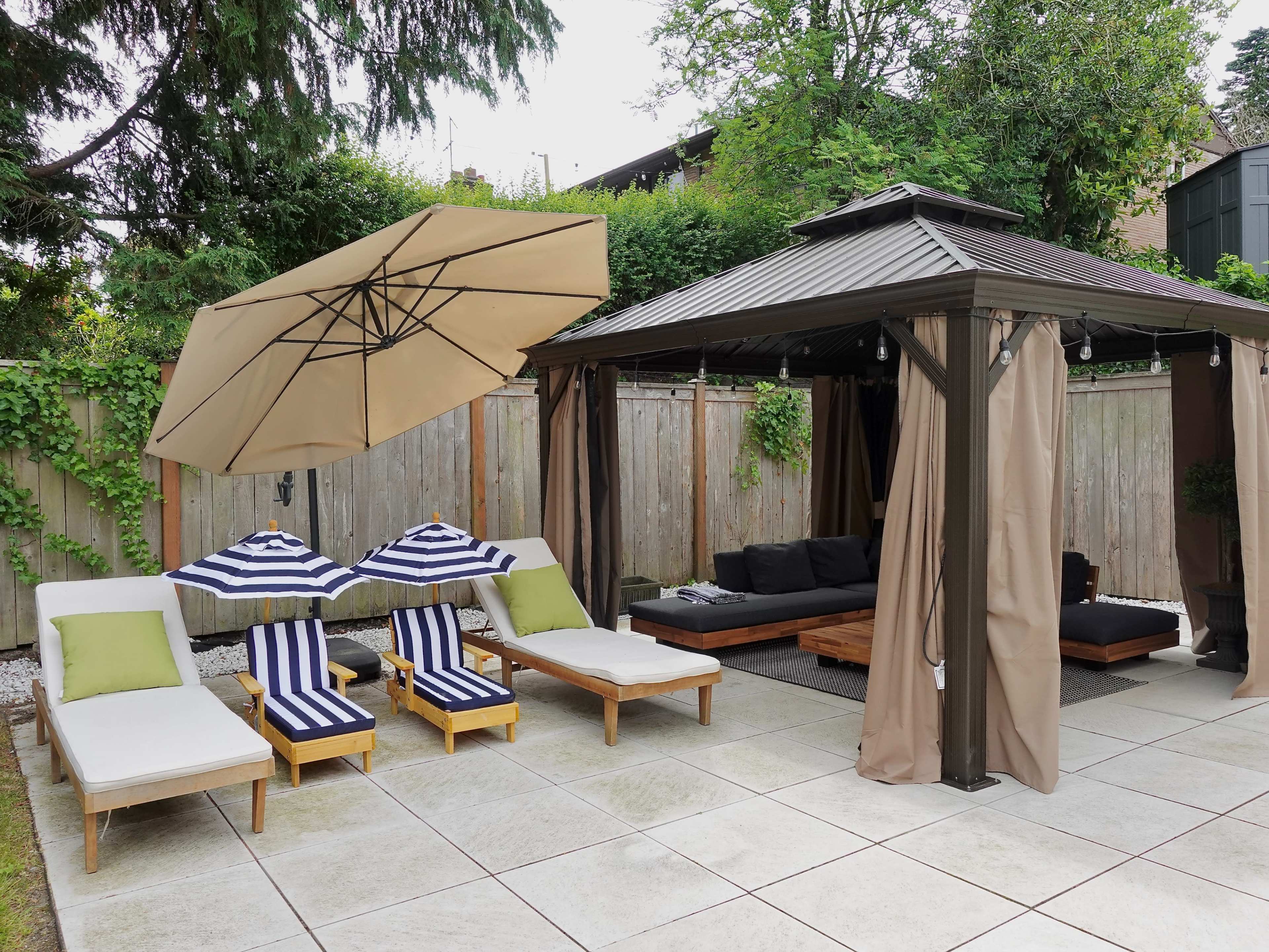 A patio area with two striped lounge chairs and an umbrella beside a gazebo with curtains, surrounded by wooden fencing and greenery.