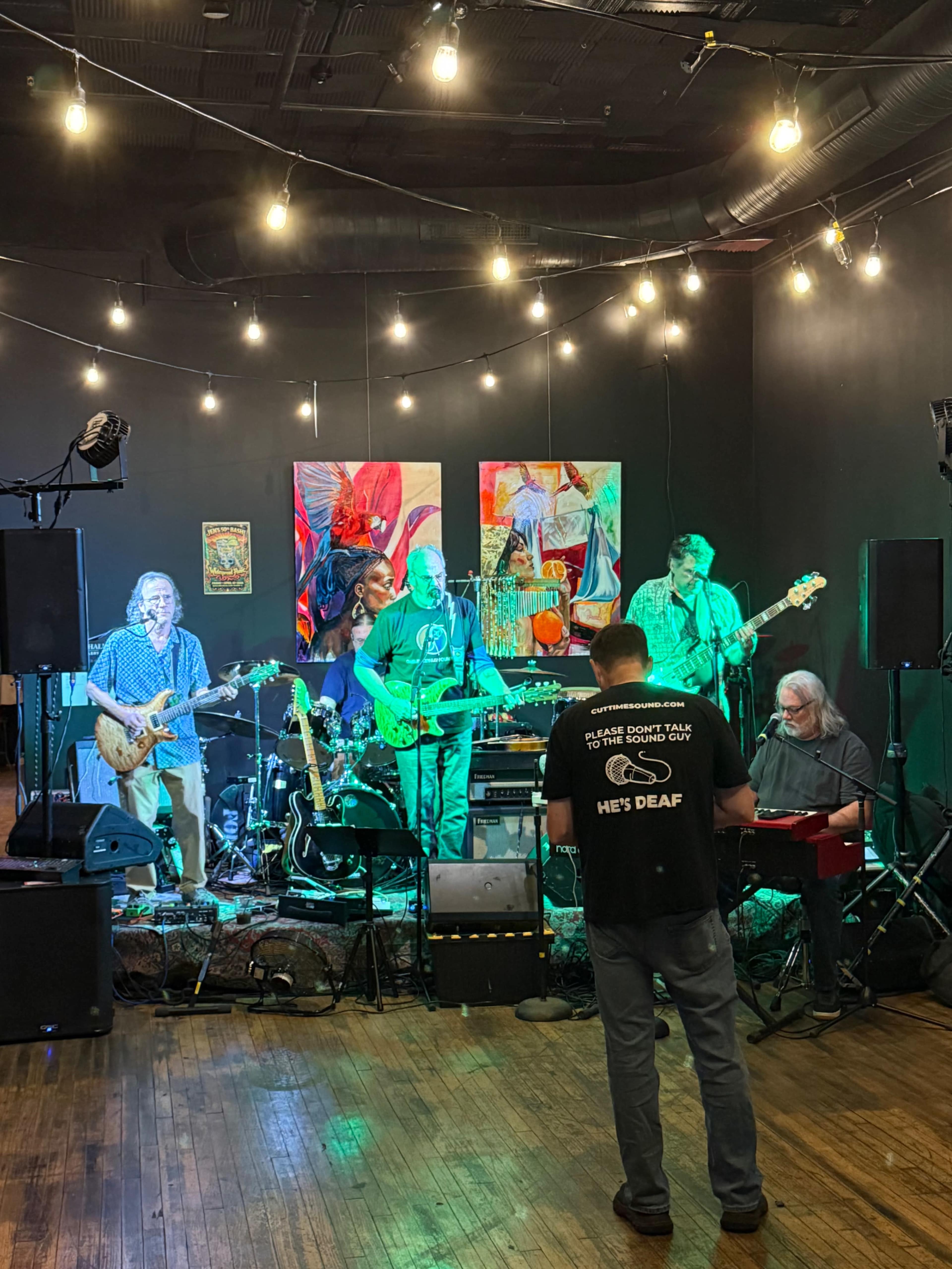A band of five musicians performs on a stage in a dimly lit venue decorated with colorful artwork and string lights.