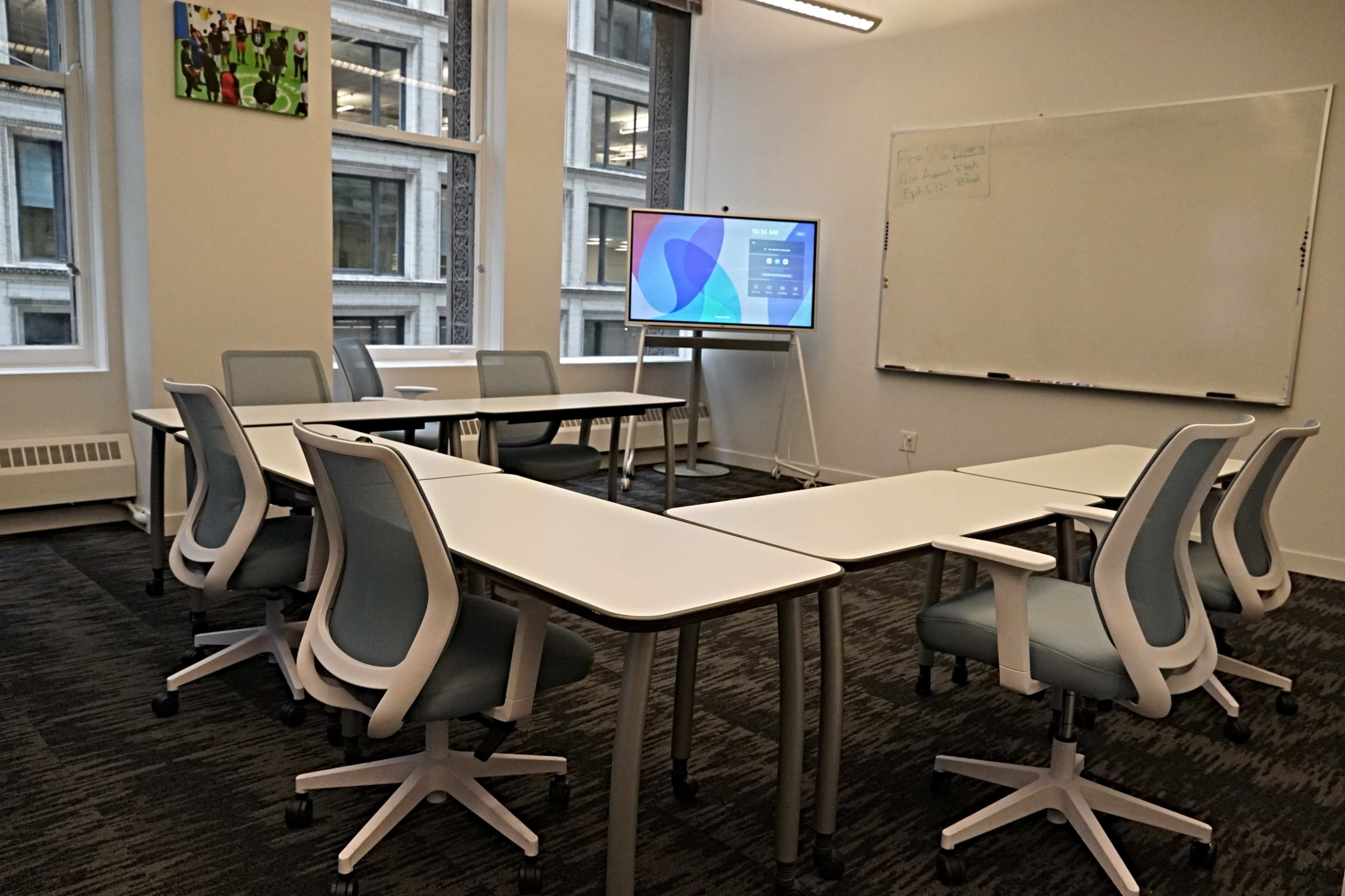 A modern classroom setup with several white tables arranged in a U-shape, accompanied by ergonomic chairs, a large screen displaying colorful graphs, and a whiteboard on the wall.