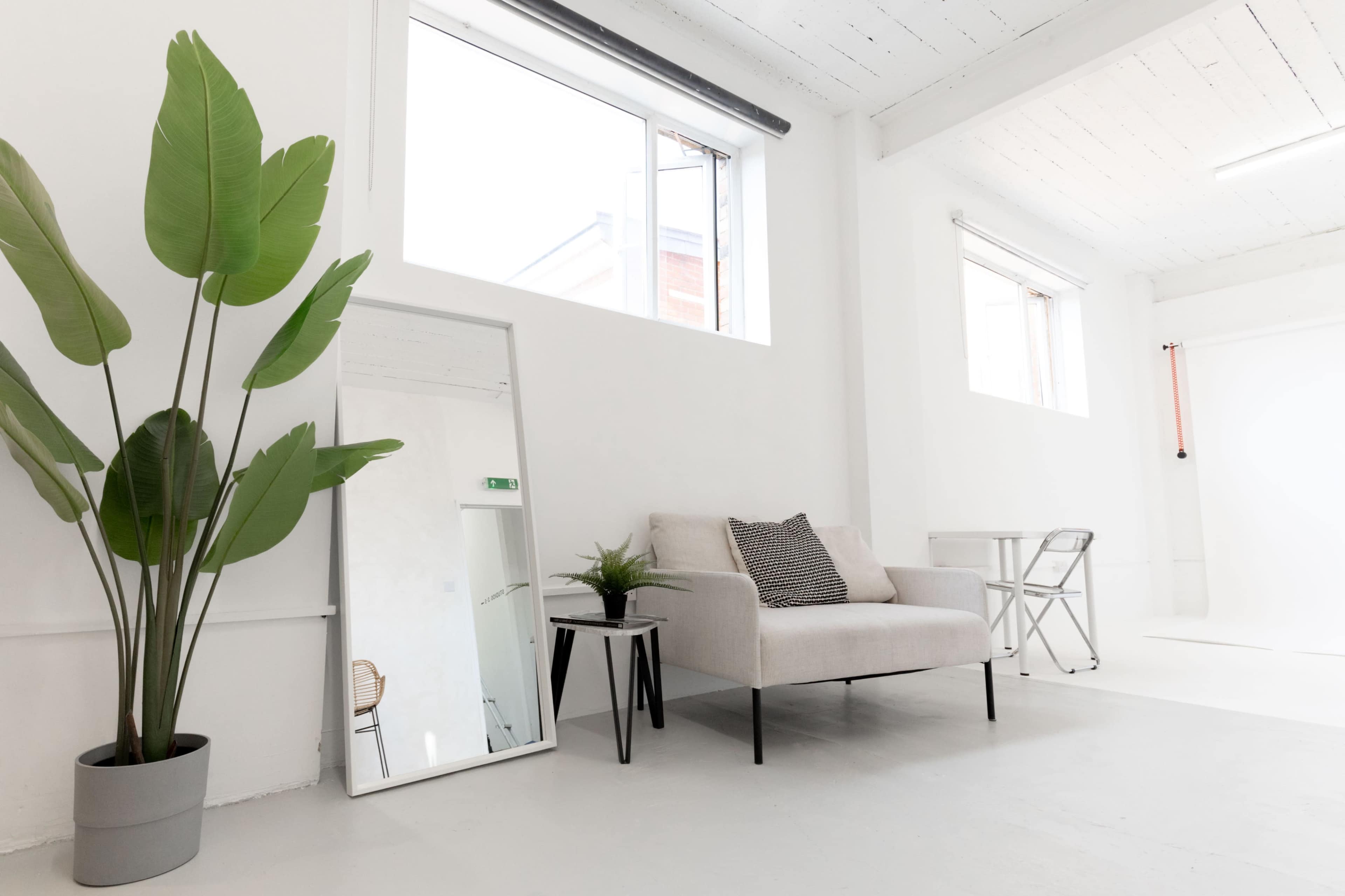 The image depicts a bright, minimalist interior space featuring a light-colored sofa, a large mirror, a small table, and a potted plant.