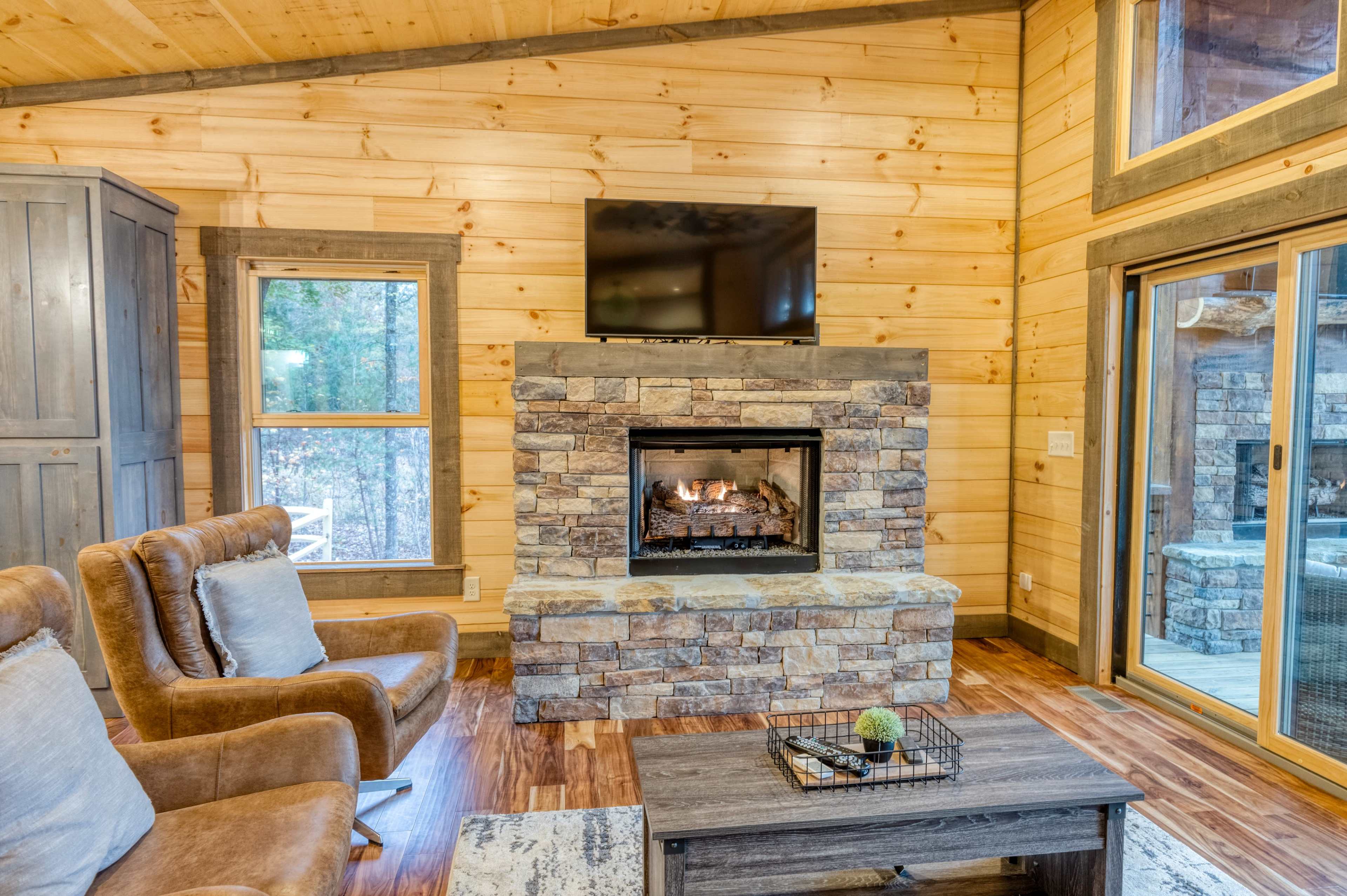 The scene features a cozy living room with a stone fireplace, a mounted television, two brown armchairs, and large windows revealing a view of the outdoors.
