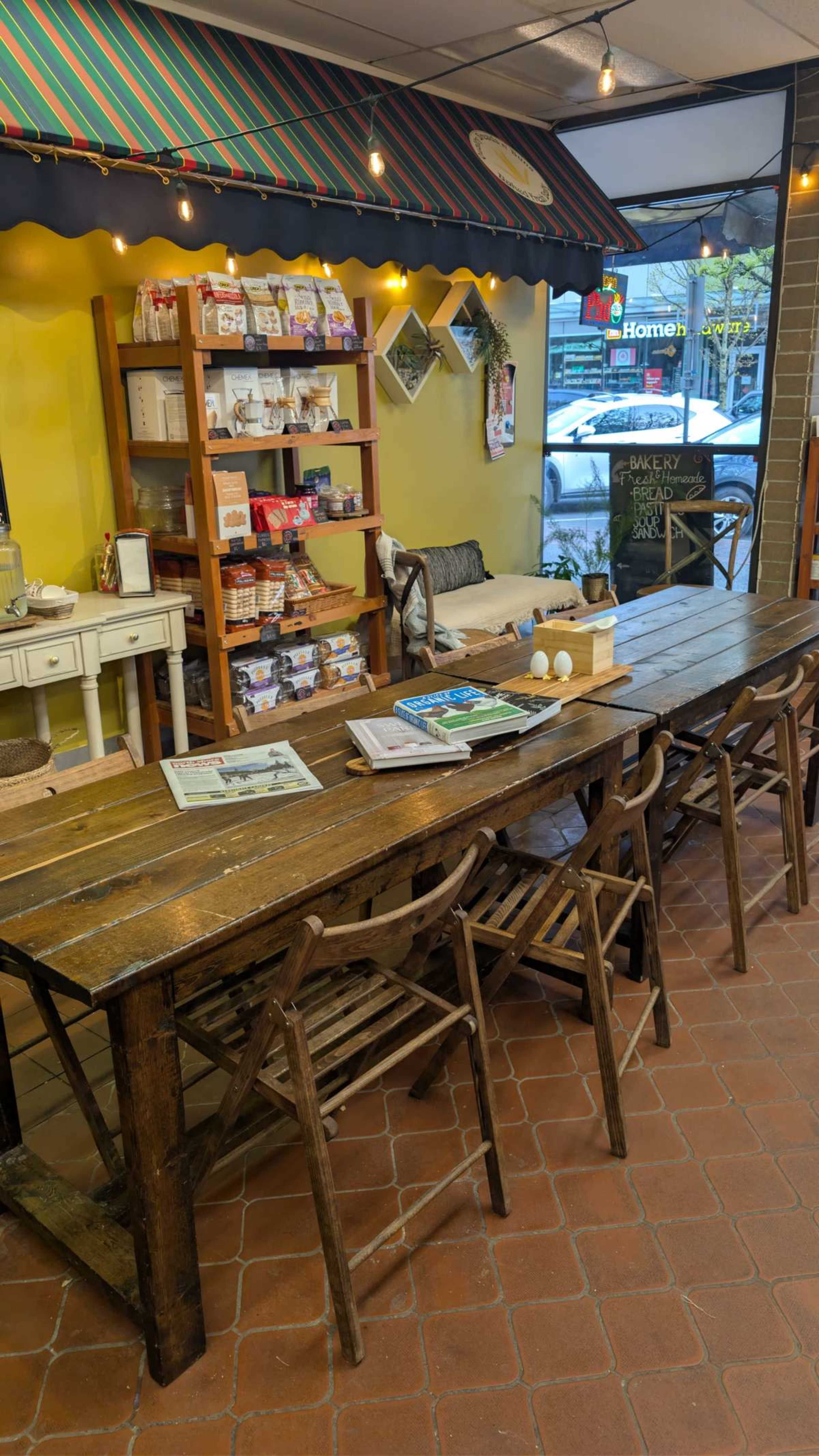 The image shows a cozy café interior featuring a wooden table surrounded by folding chairs, with a colorful display of products on shelves against a yellow wall.