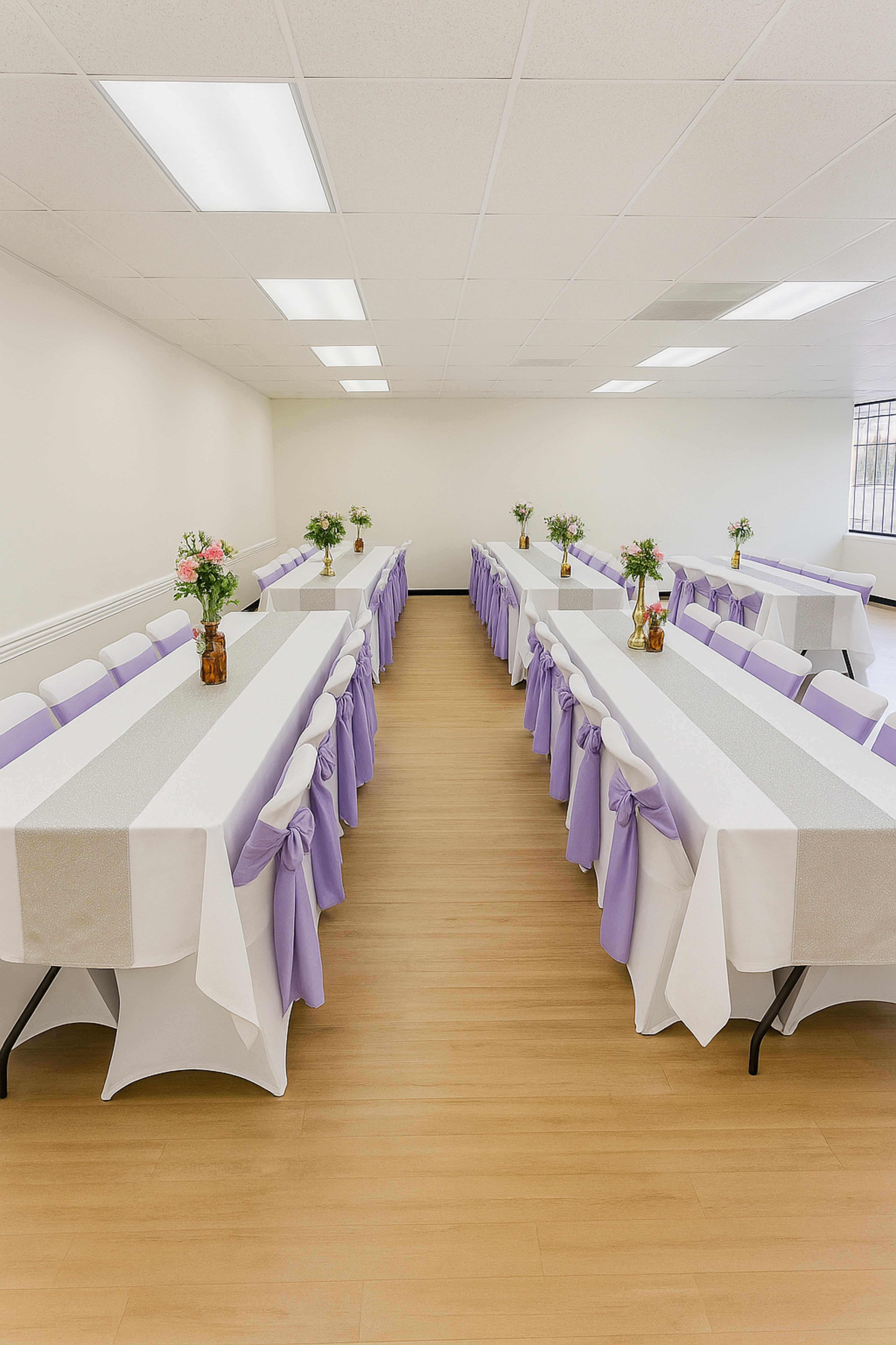The image shows a spacious interior with two long tables arranged in a U-shape, decorated with lavender sashes and vases of flowers on each table.