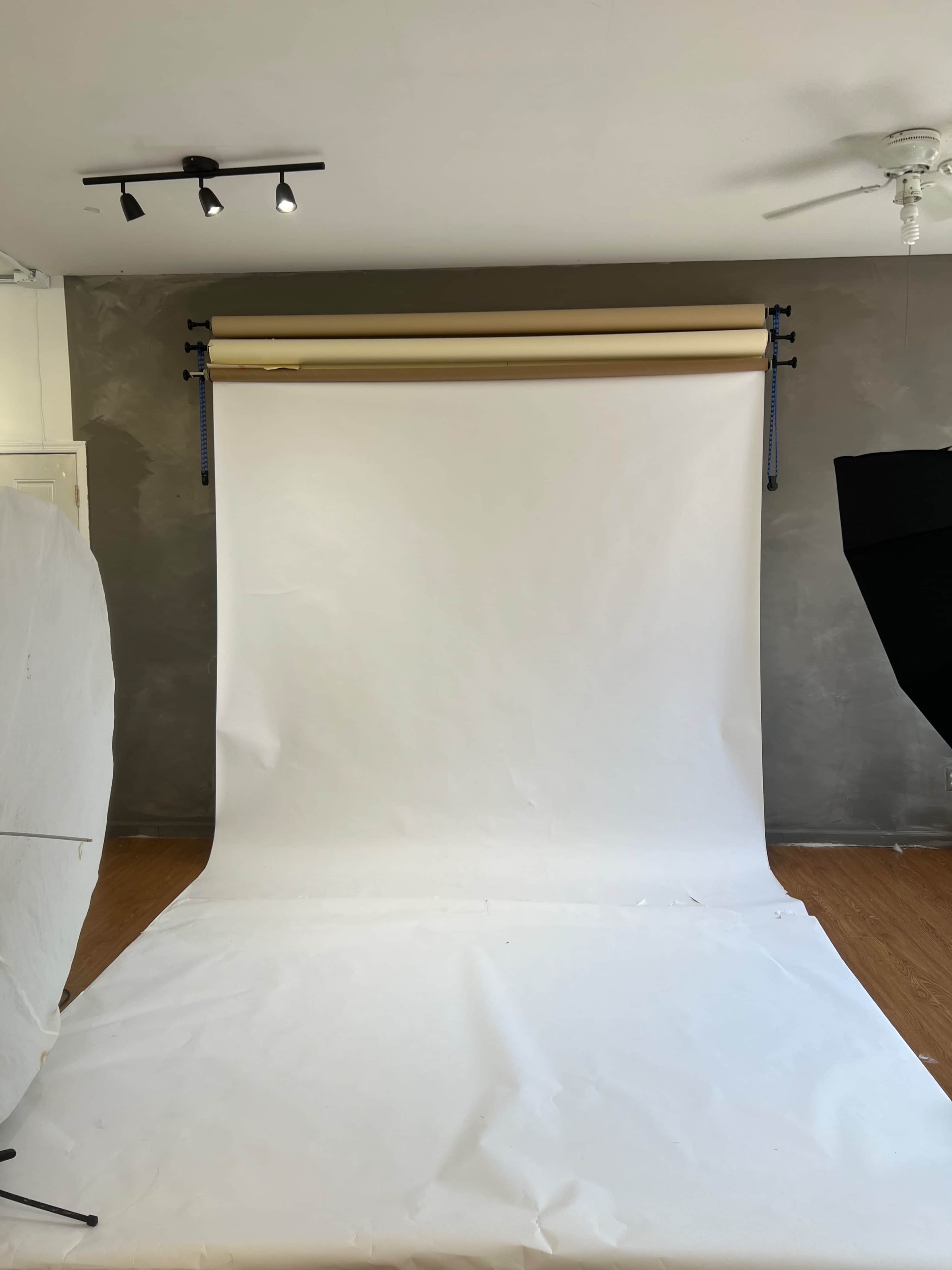 The image shows a photography studio with a large white backdrop, lighting equipment, and a wooden floor.