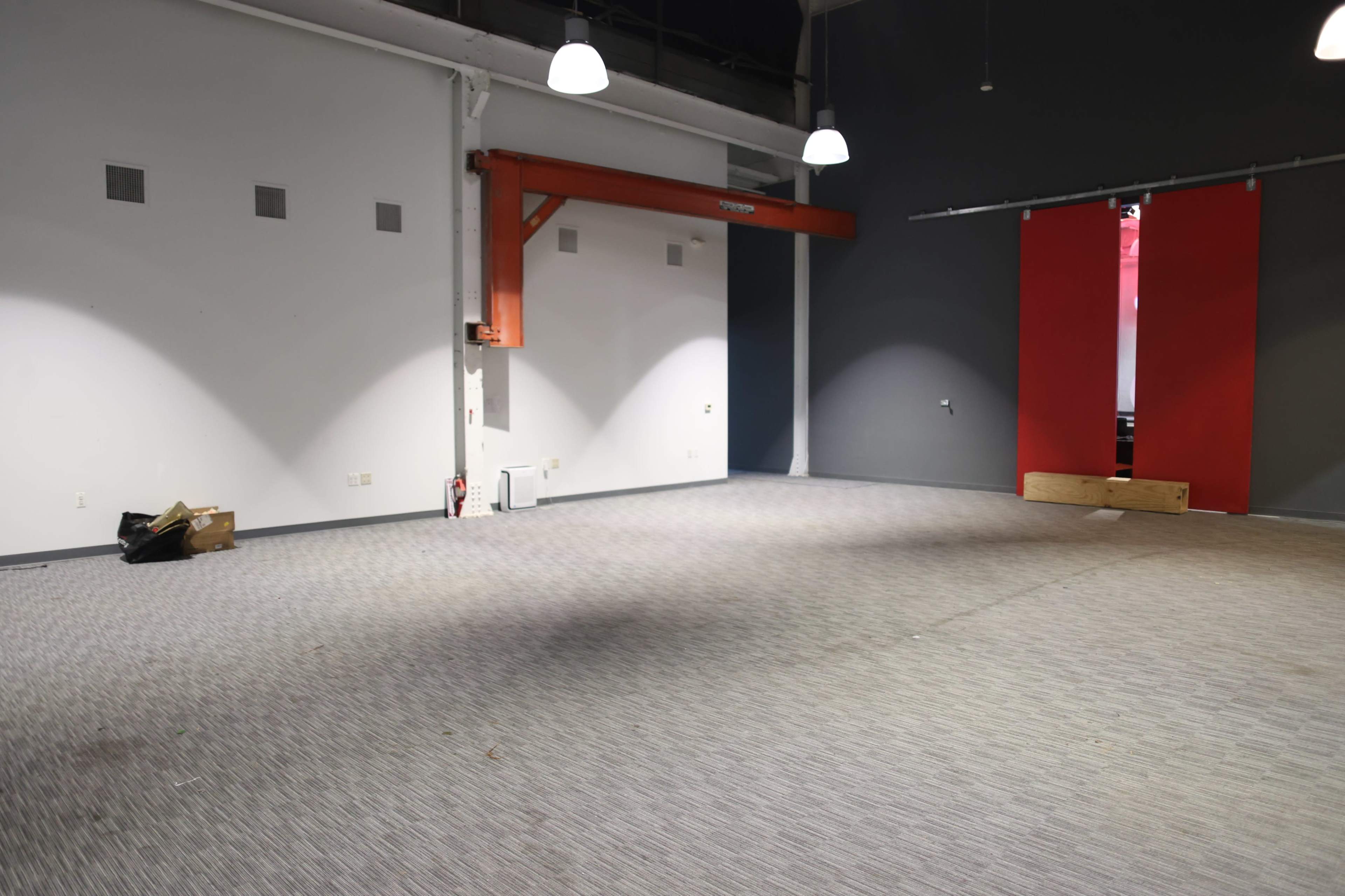 The image shows an empty, spacious room with gray walls, a light gray carpet, and a red accent wall, featuring a set of closed doors.