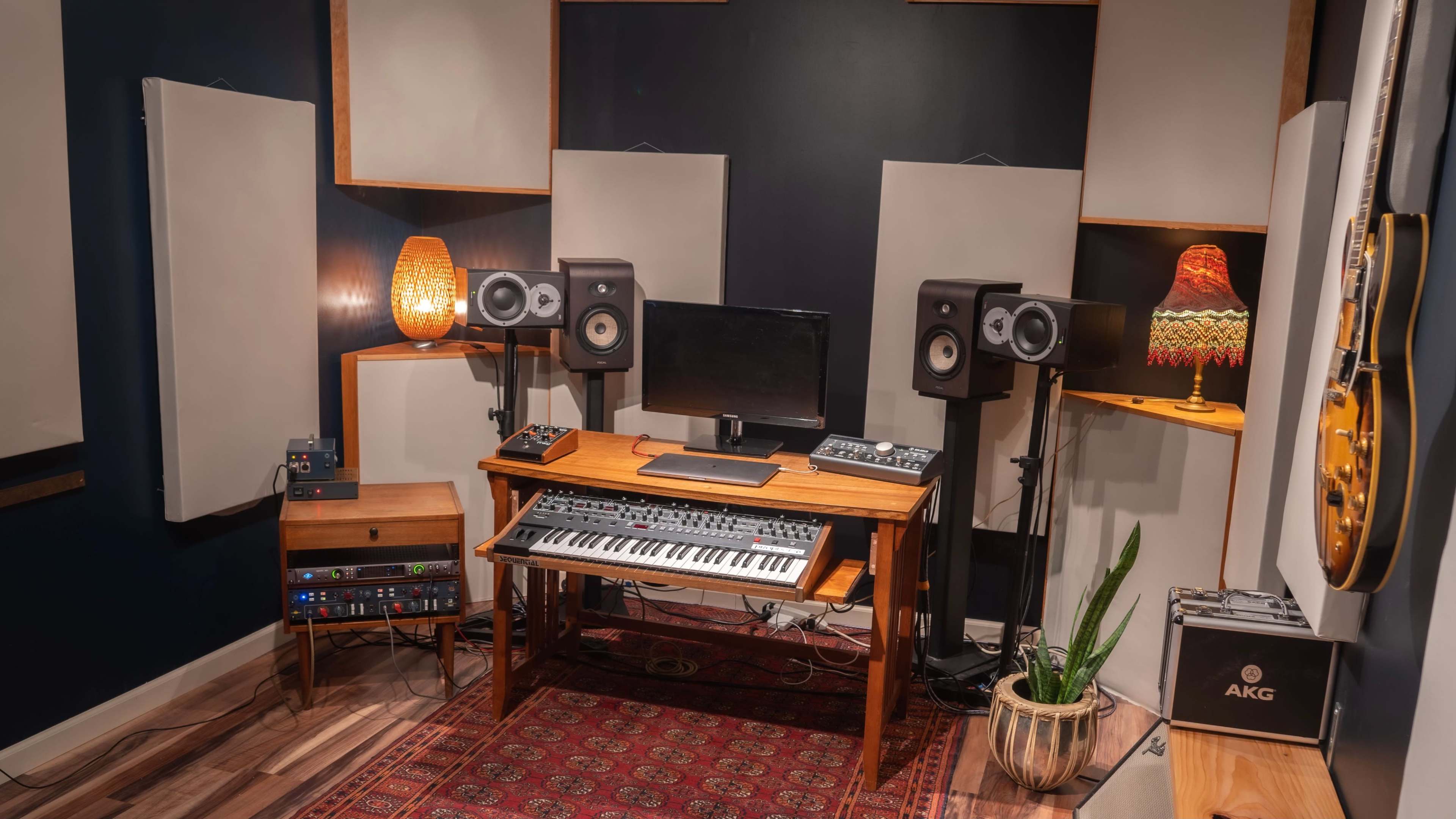 A music studio setup featuring a wooden desk with a keyboard and computer monitor flanked by two speakers, acoustic panels on the walls, and a guitar on display.