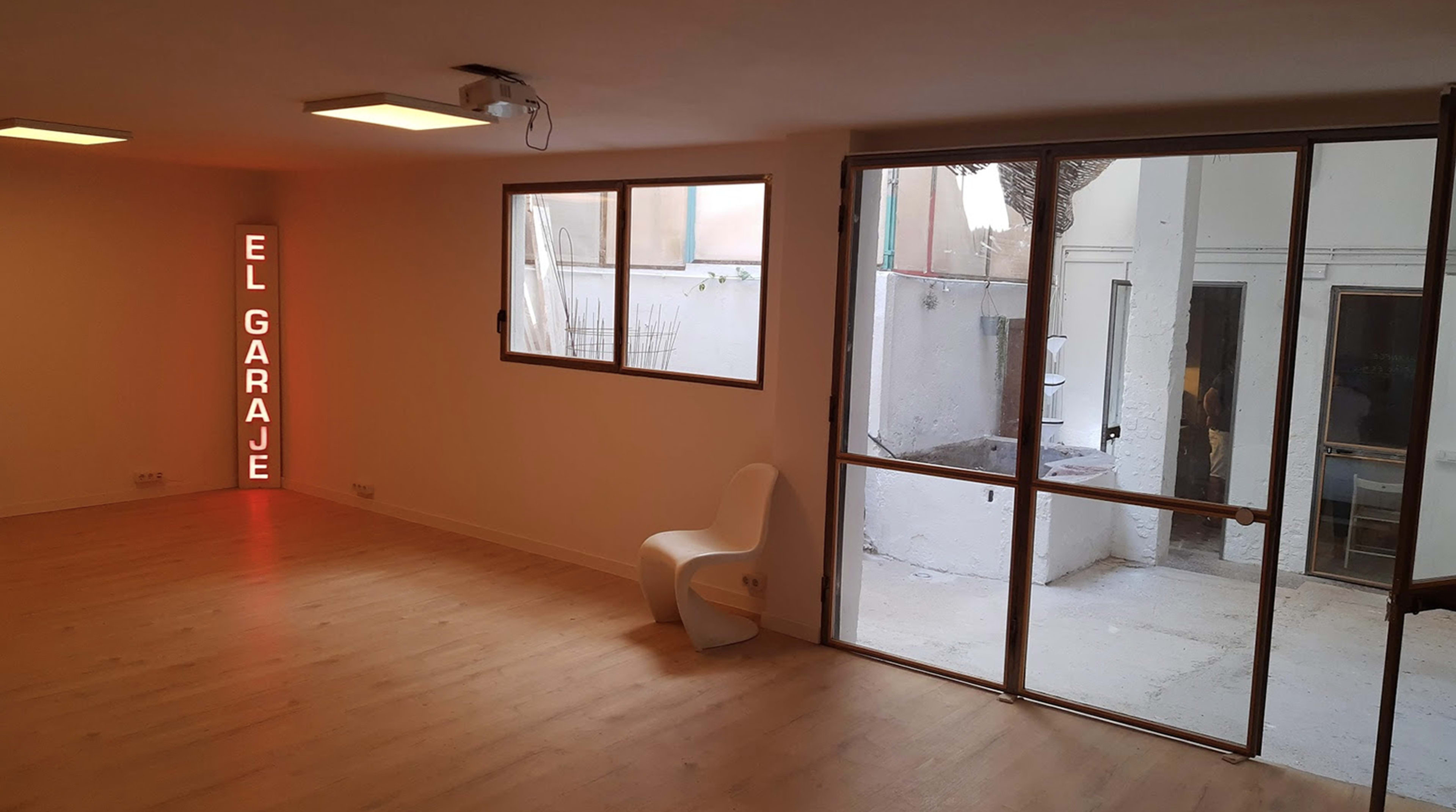 The image shows a minimalist room featuring a neon sign that reads "EL GARAGE," a white chair, and large glass doors leading to an outdoor area.