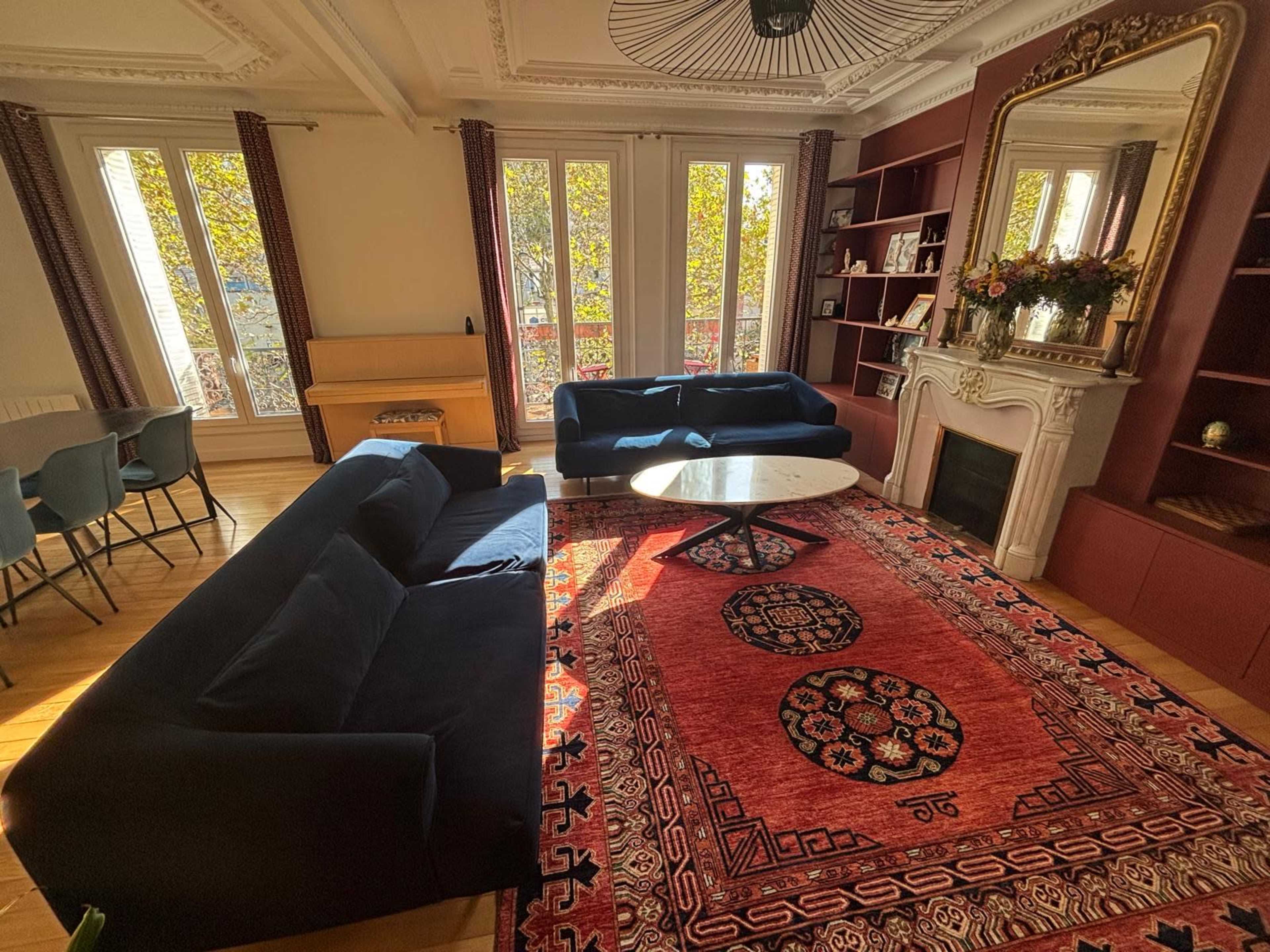 The image shows a spacious living room featuring two dark sofas, a round coffee table, a patterned area rug, and large windows with leafy views.