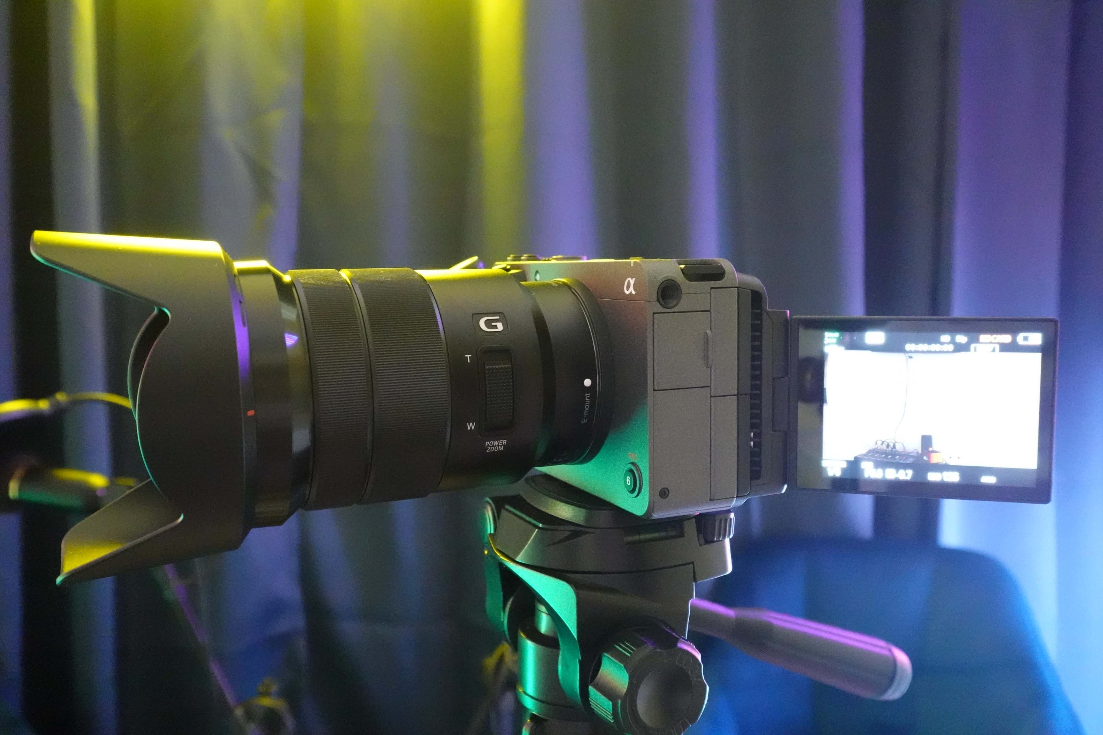 A camera with a lens and viewfinder is positioned on a tripod against a backdrop of dark fabric illuminated by colored lights.