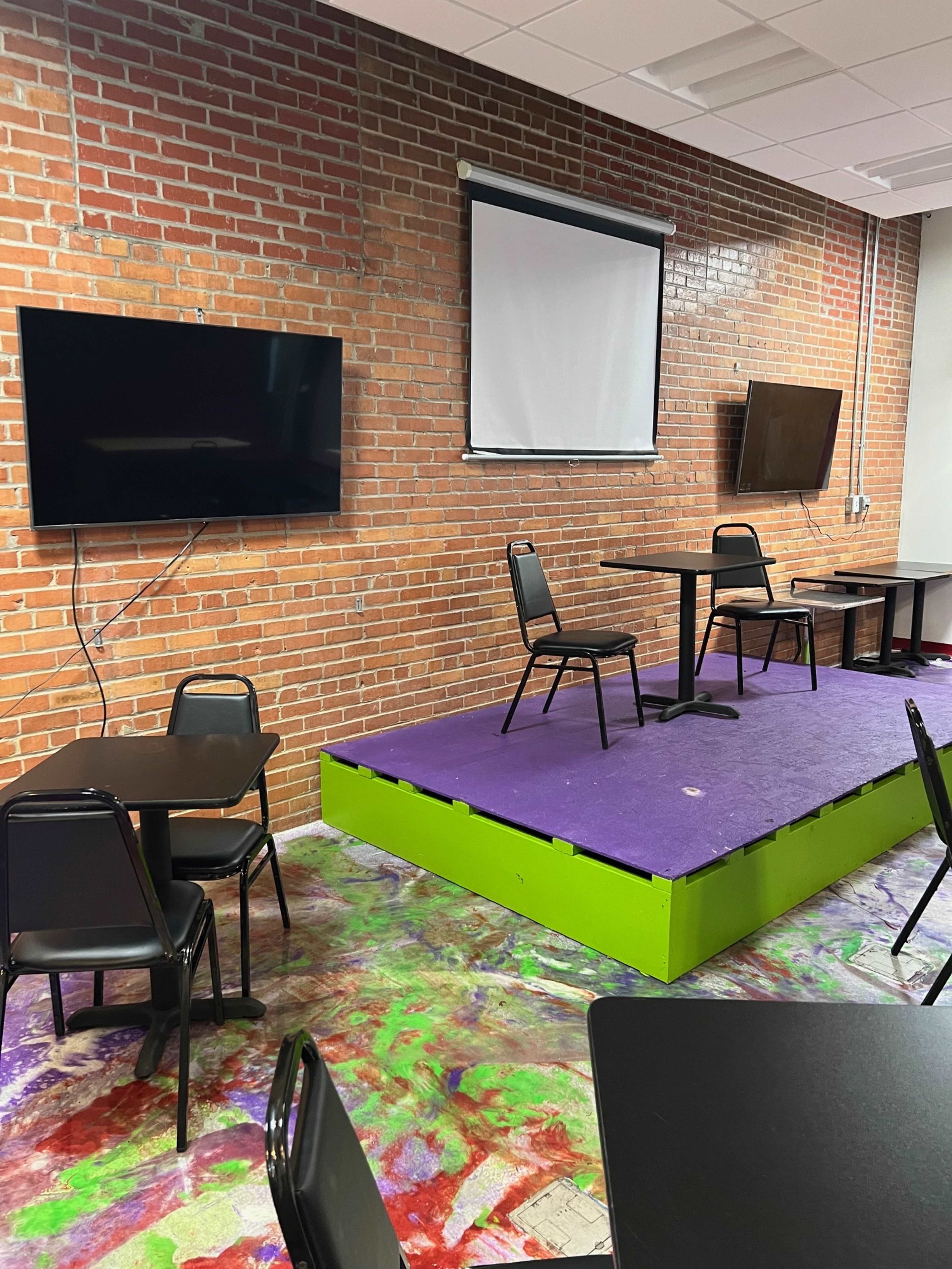 A small room with brick walls, two flat-screen TVs mounted on the wall, a raised platform in the center, and several black chairs and tables arranged around it.