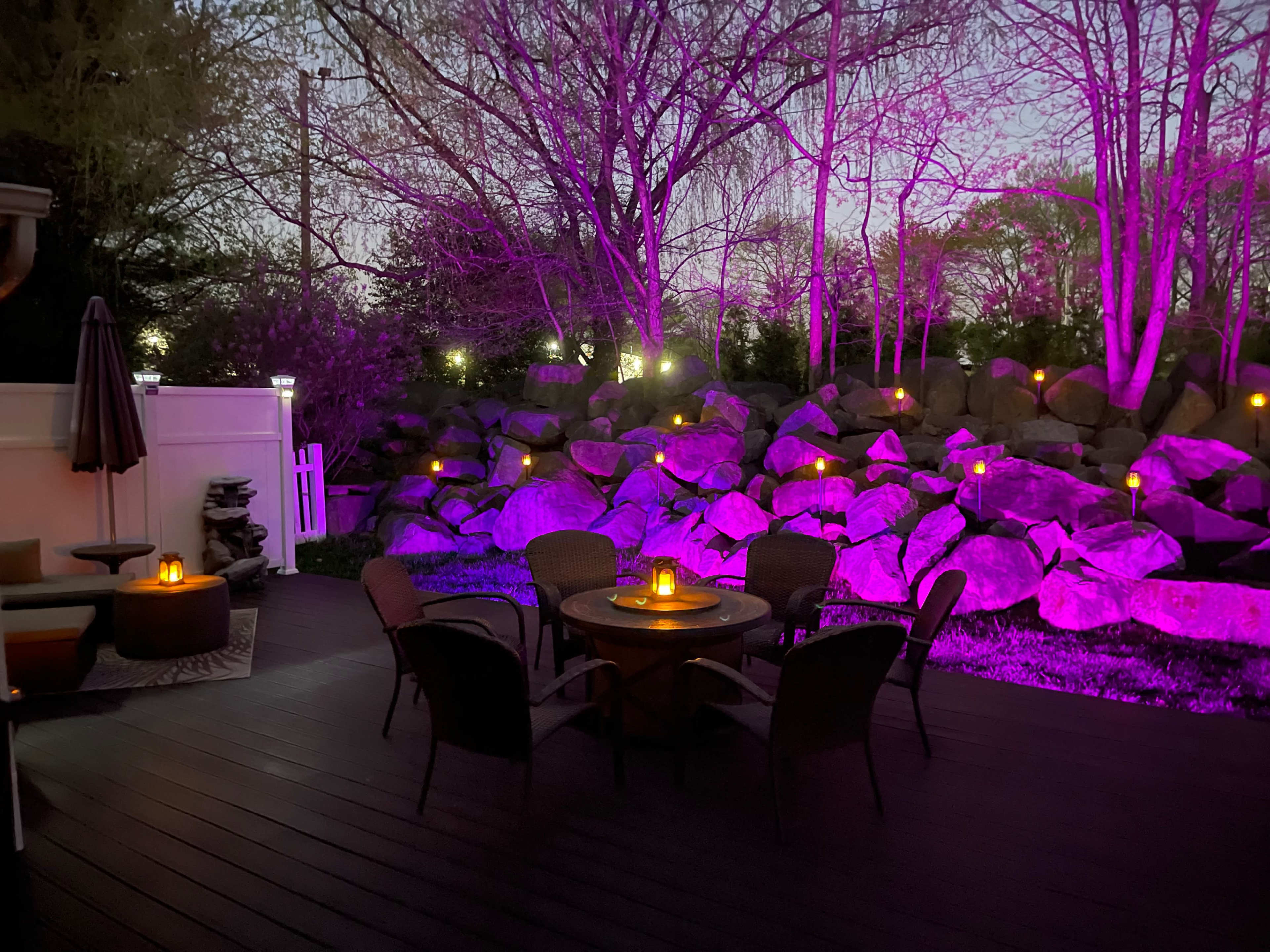 A seating area with circular table and chairs is illuminated by candles and purple lights near a rock garden at dusk.