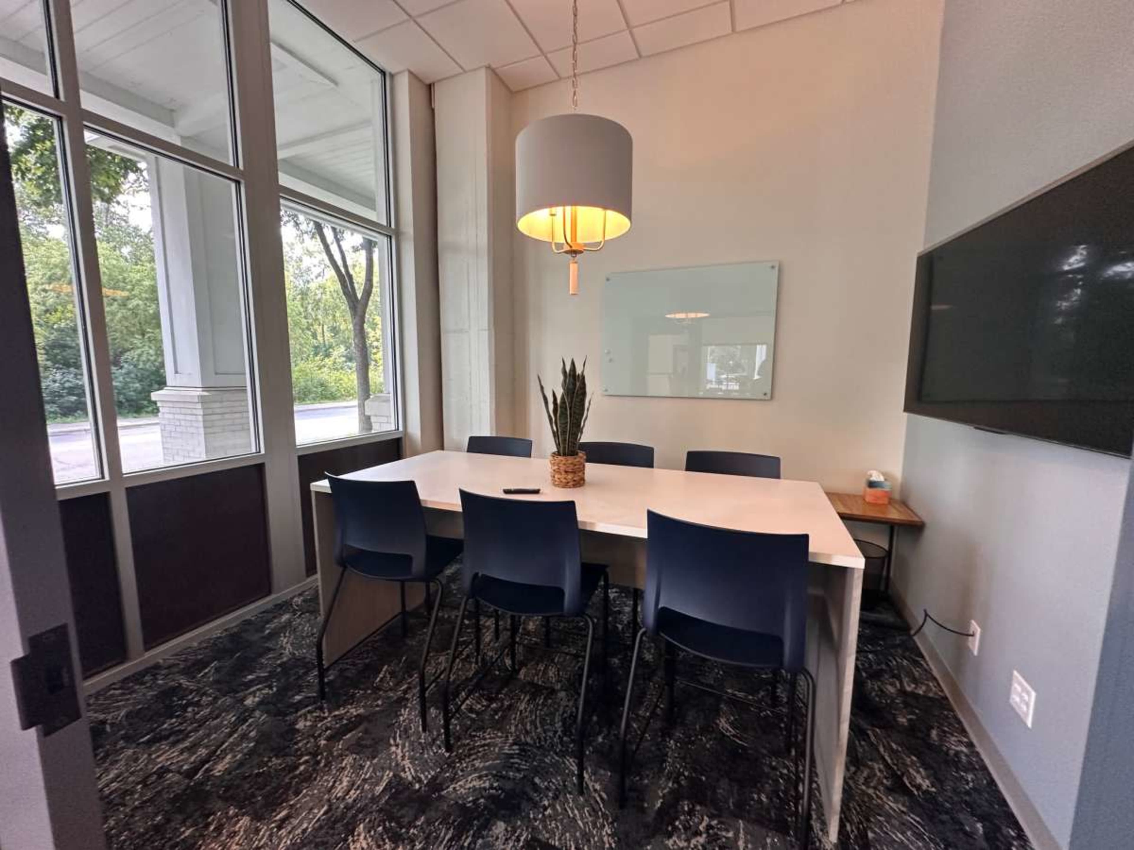 A modern conference room features a large table surrounded by chairs, a wall-mounted screen, and a large window offering a view of greenery outside.