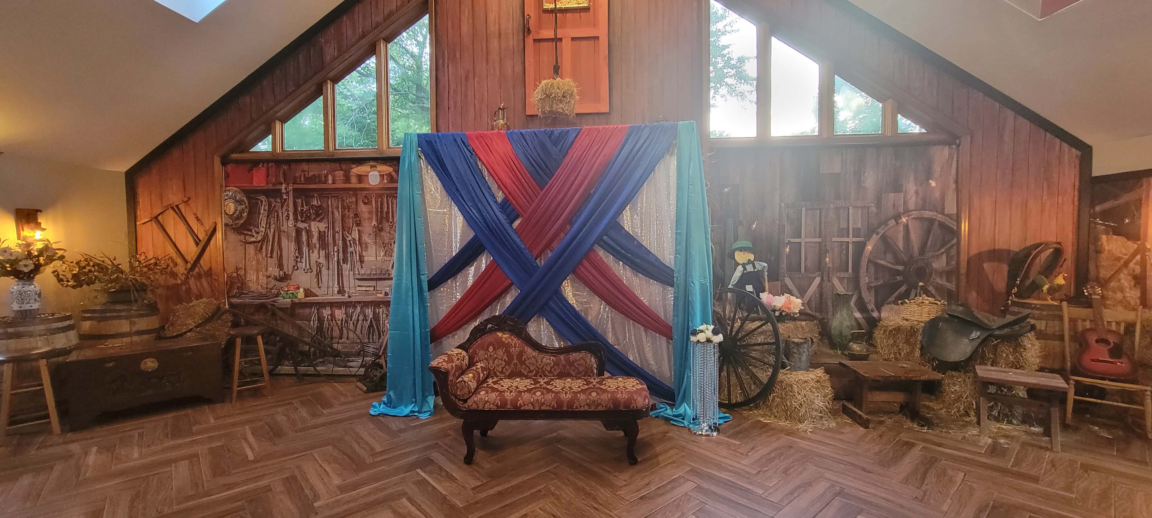 A decorated interior with a backdrop of draped fabric in red and blue, a vintage couch, and rustic decor elements including barrels, hay, and farming tools.