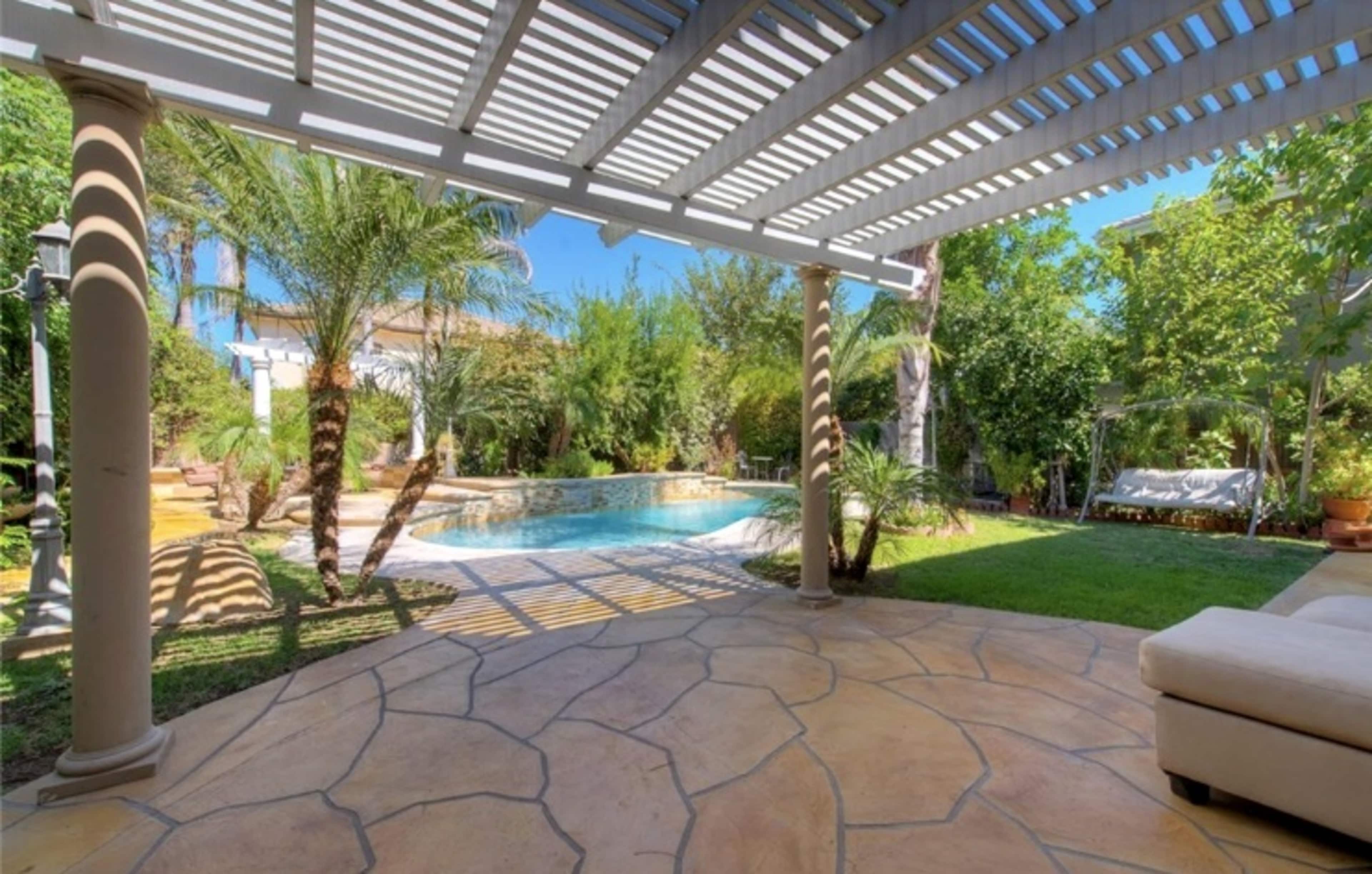 The image shows a backyard with a swimming pool surrounded by greenery and shaded by a pergola.