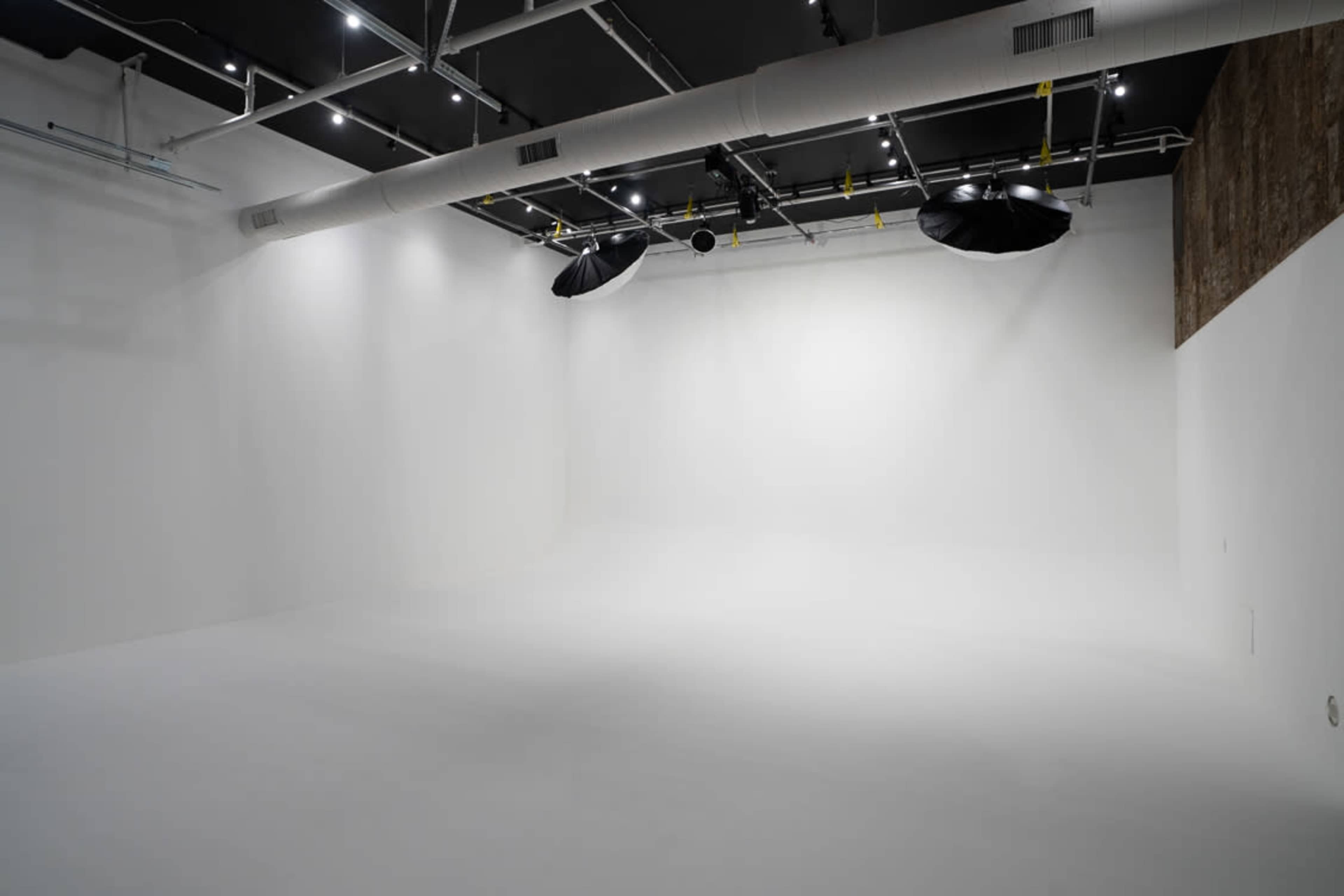 The image shows a spacious studio with white walls, a white floor, and two studio lights hanging from the ceiling.