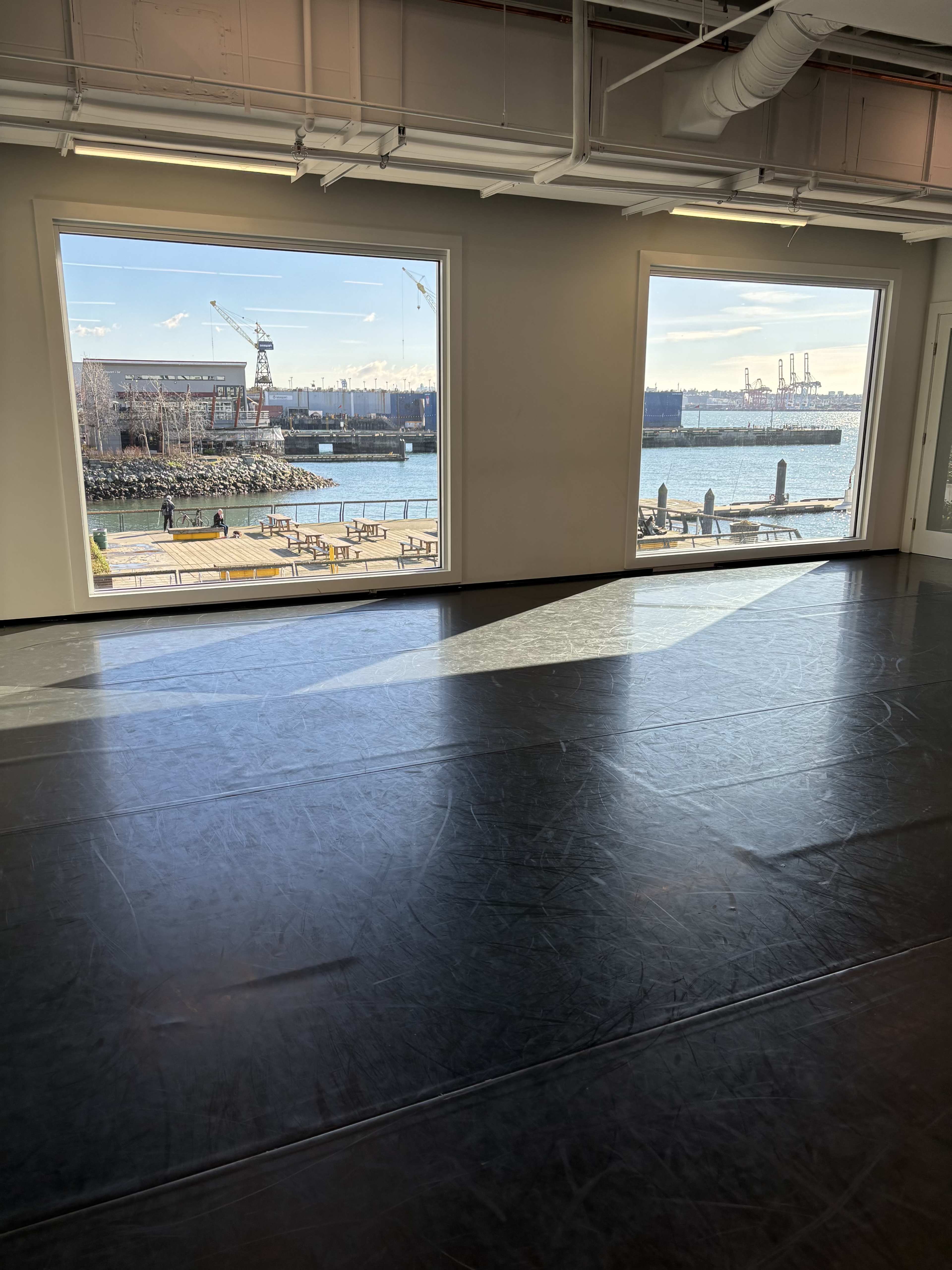 The image shows an indoor space with large windows that overlook a body of water and a dock area, with benches and people visible outside.