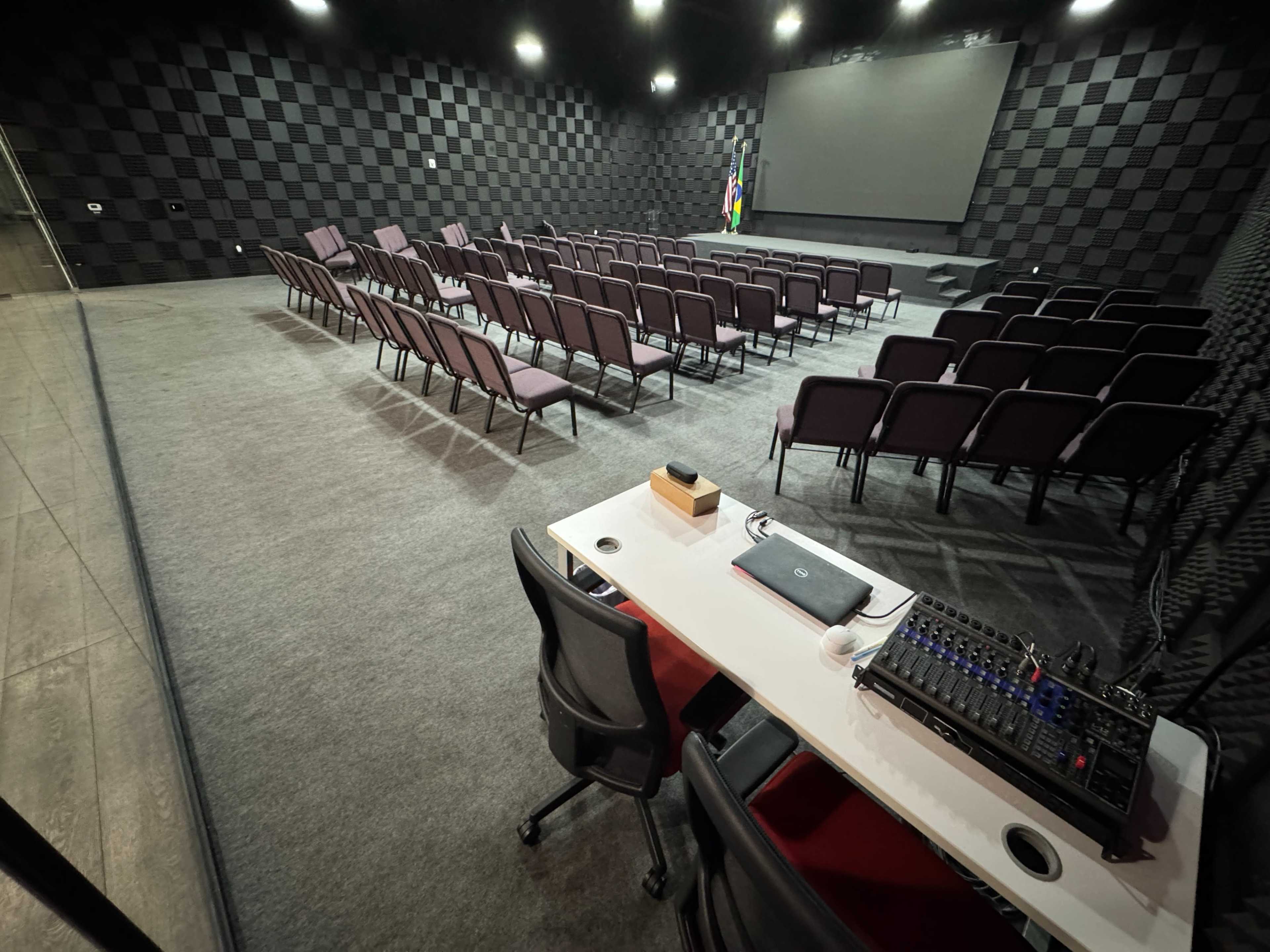 The image shows a theater-like room with rows of purple seating facing a large blank screen, accompanied by a sound mixing station in the foreground.