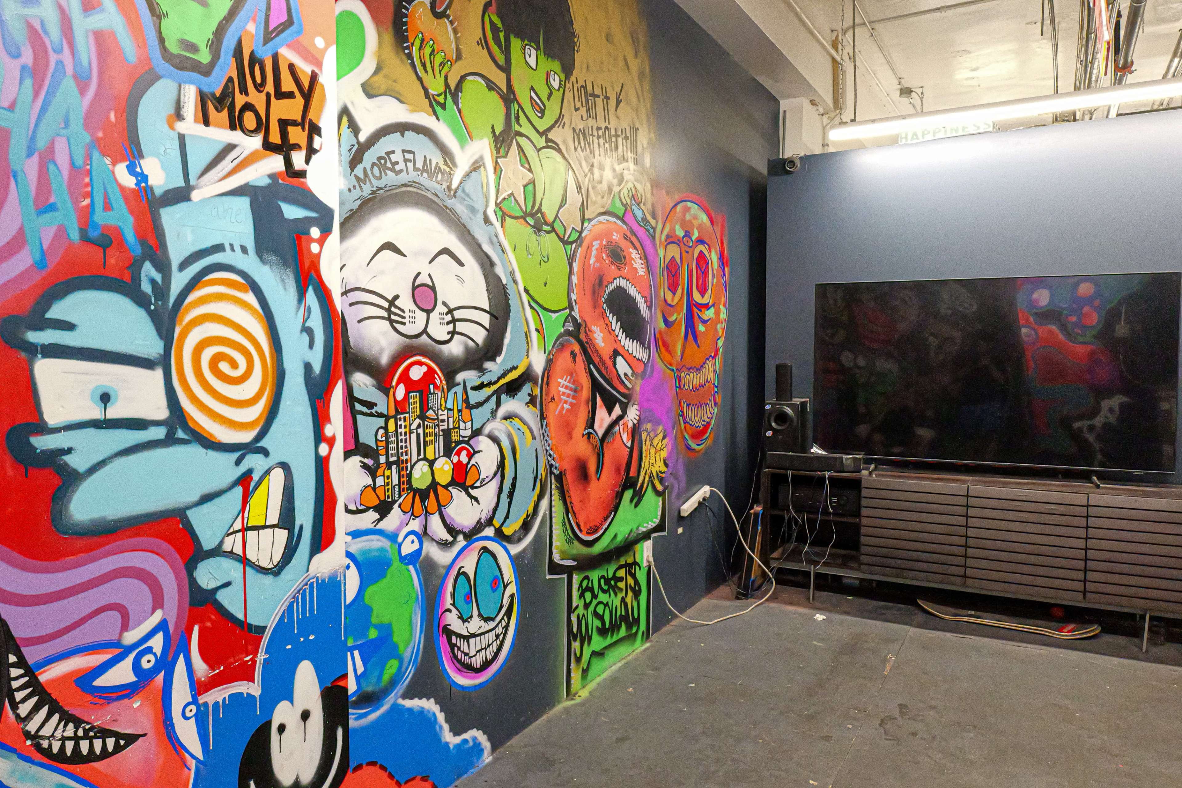 A colorful wall covered in graffiti art is adjacent to a modern television set and a low media console in a dimly lit room.