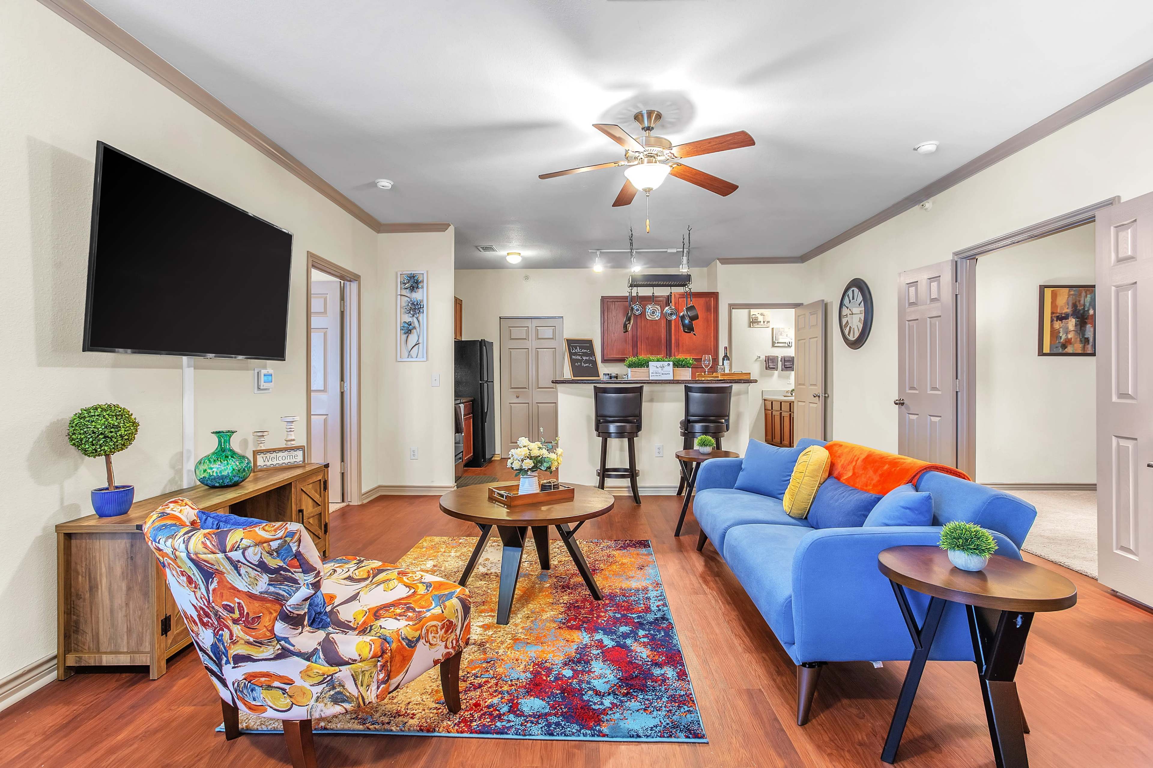 The image shows a living room with a blue sofa, an armchair with a colorful pattern, a round coffee table, and a dining area in the background featuring bar stools.