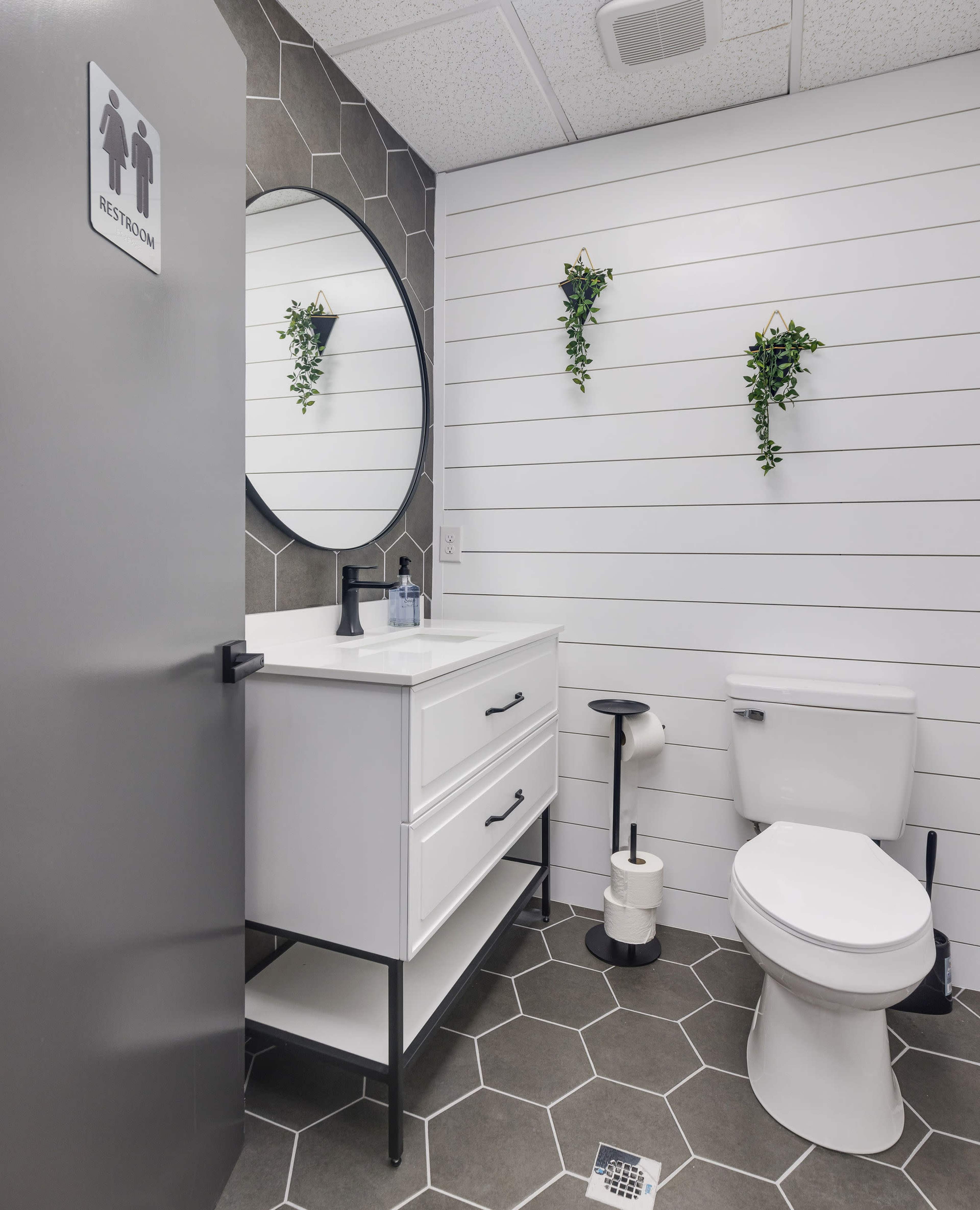 A modern bathroom with a round mirror, a white vanity with drawers, a toilet, and hexagonal floor tiles, accented by two hanging plants on the wall.