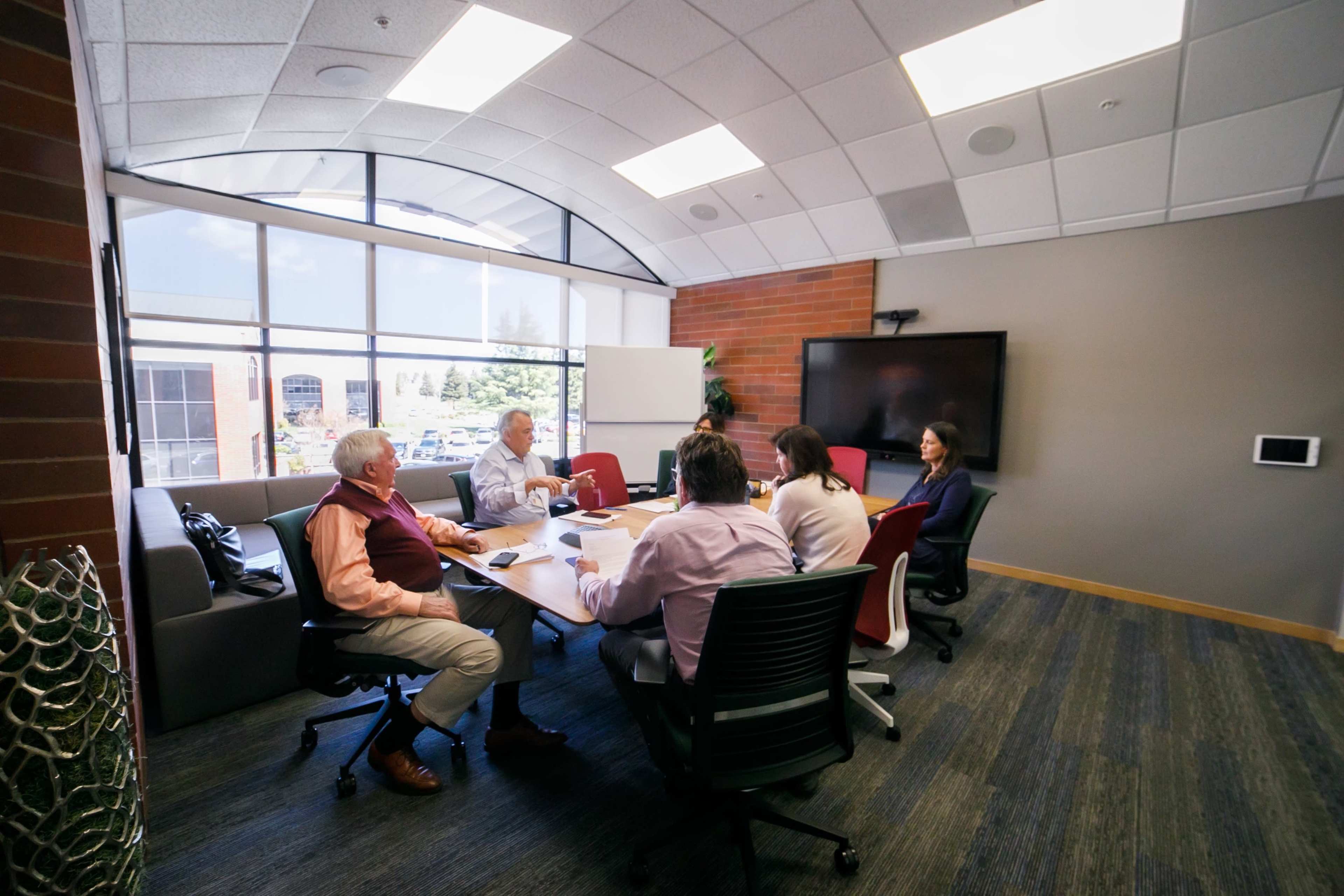 A group of five people is seated around a conference table in a brightly lit meeting room with large windows and modern furnishings.