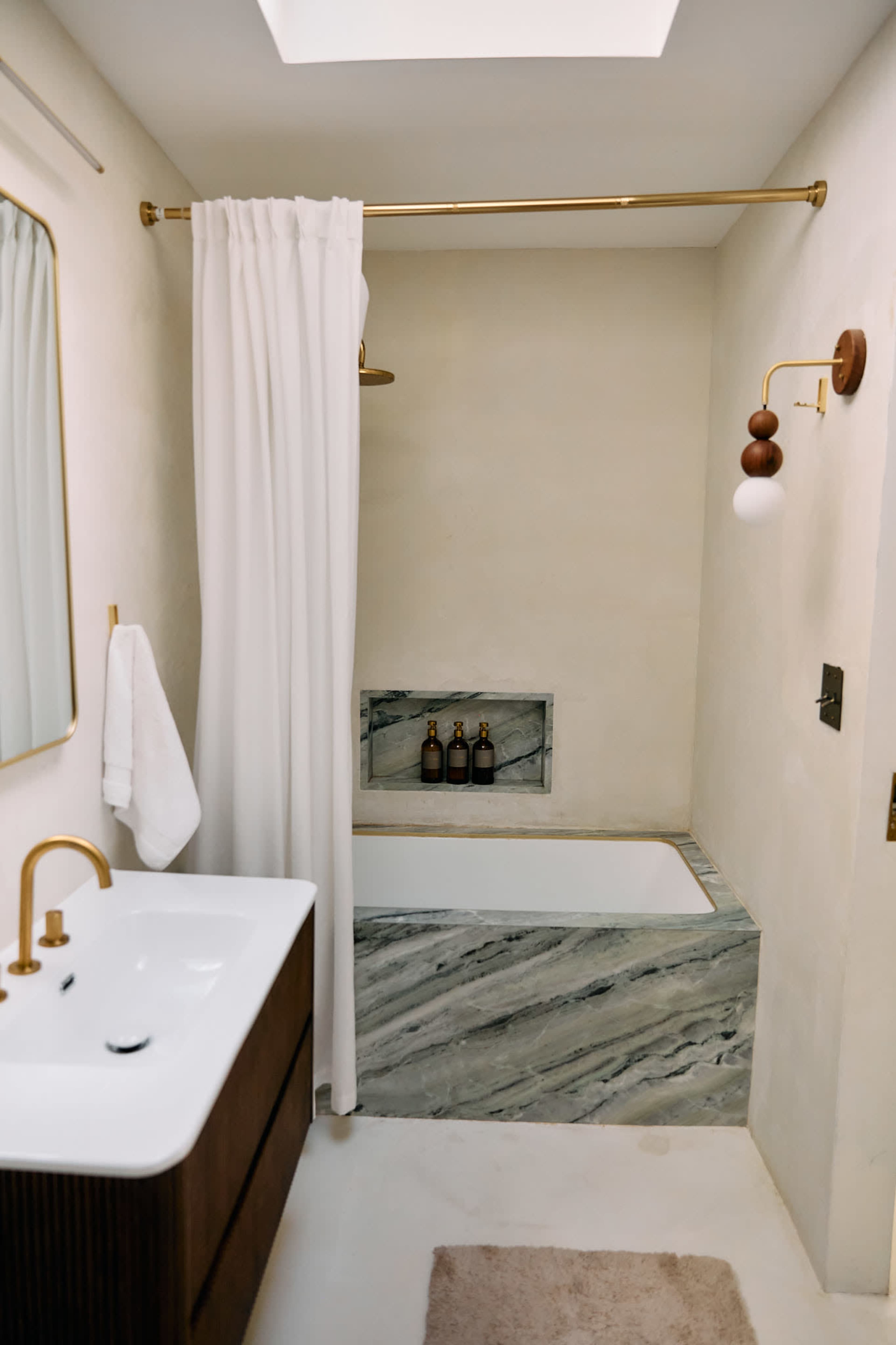 The image shows a modern bathroom featuring a freestanding bathtub, a minimalist vanity with a sink, a shower with a curtain, and a skylight above.