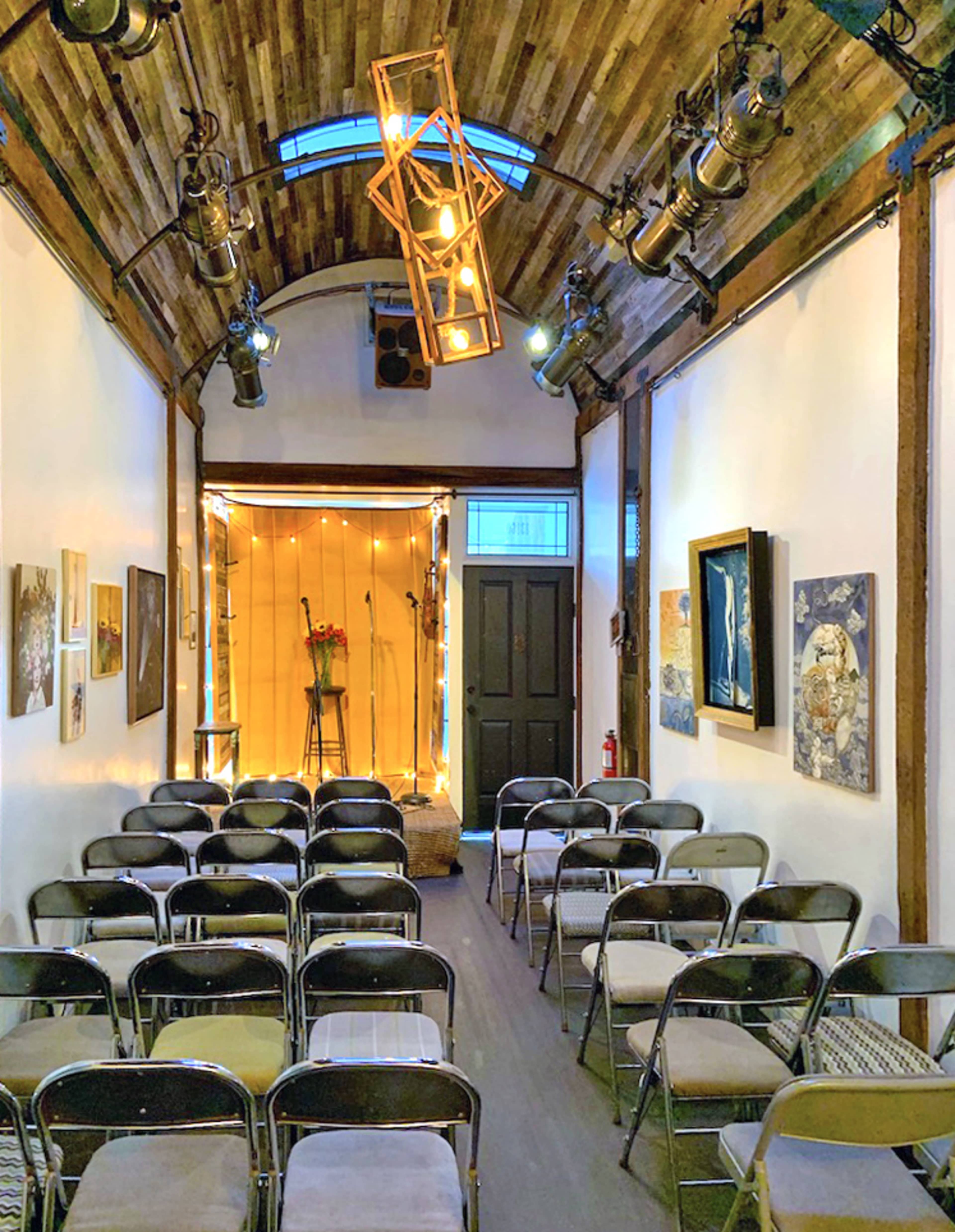 A small performance space with rows of metal chairs facing a stage adorned with a flower arrangement and string lights.