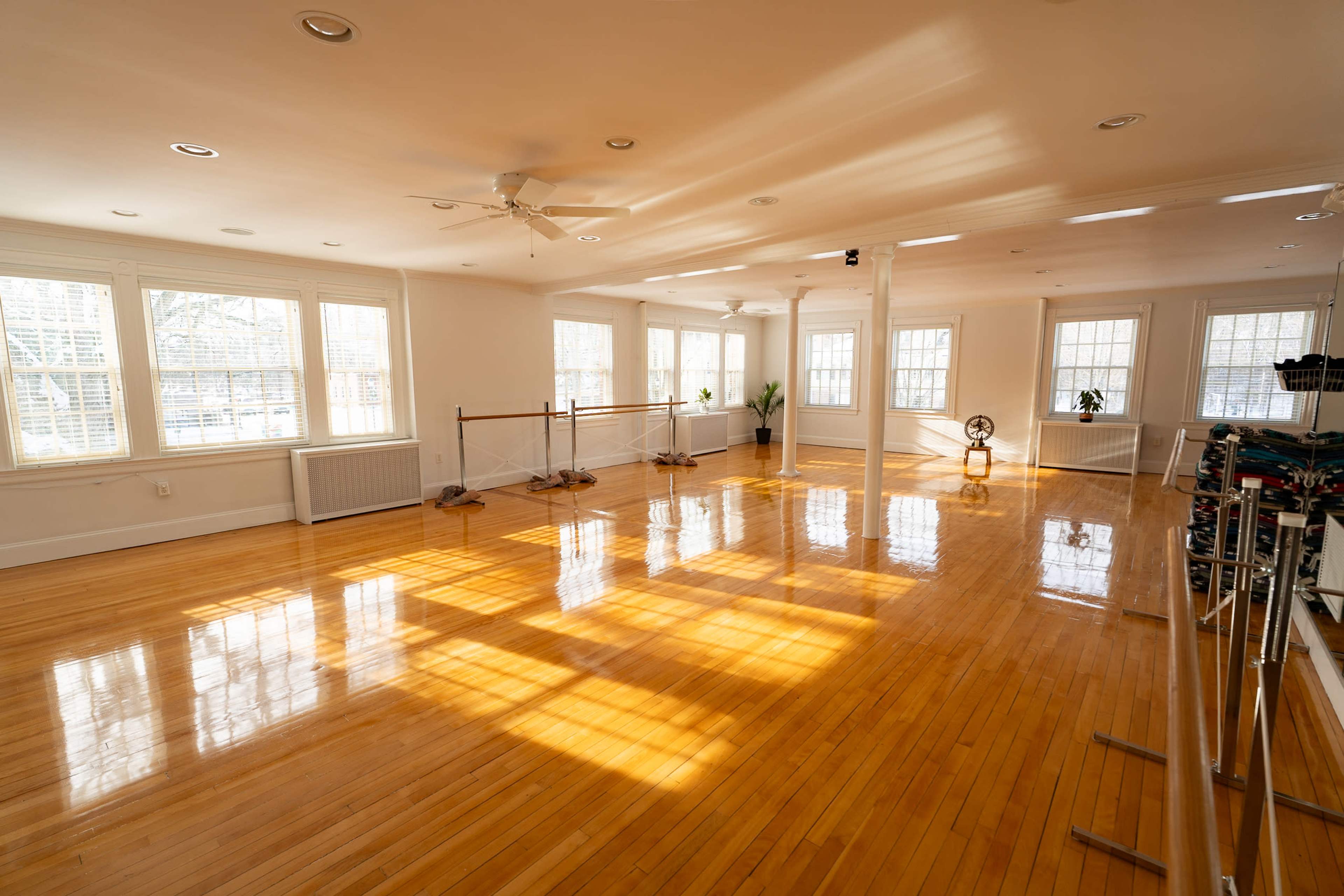 The image shows a spacious, well-lit dance studio with polished wooden floors and ballet barres along one wall.