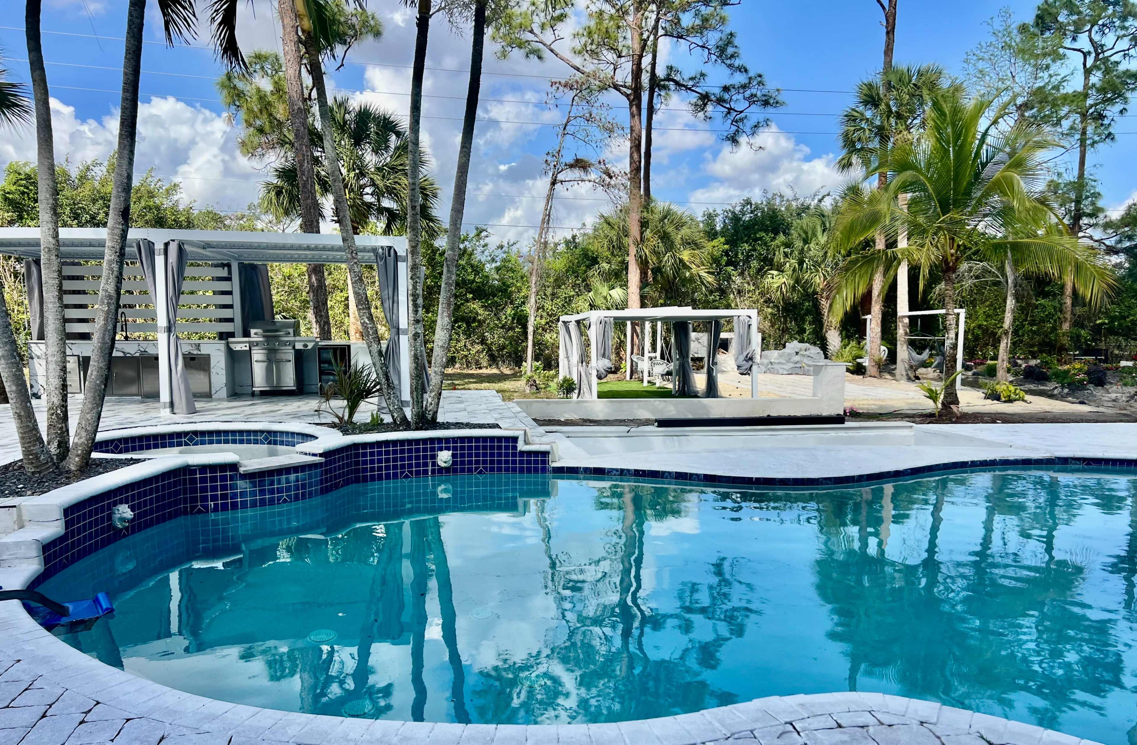 A modern backyard features a swimming pool surrounded by palm trees, with a shaded outdoor kitchen and seating area visible nearby.