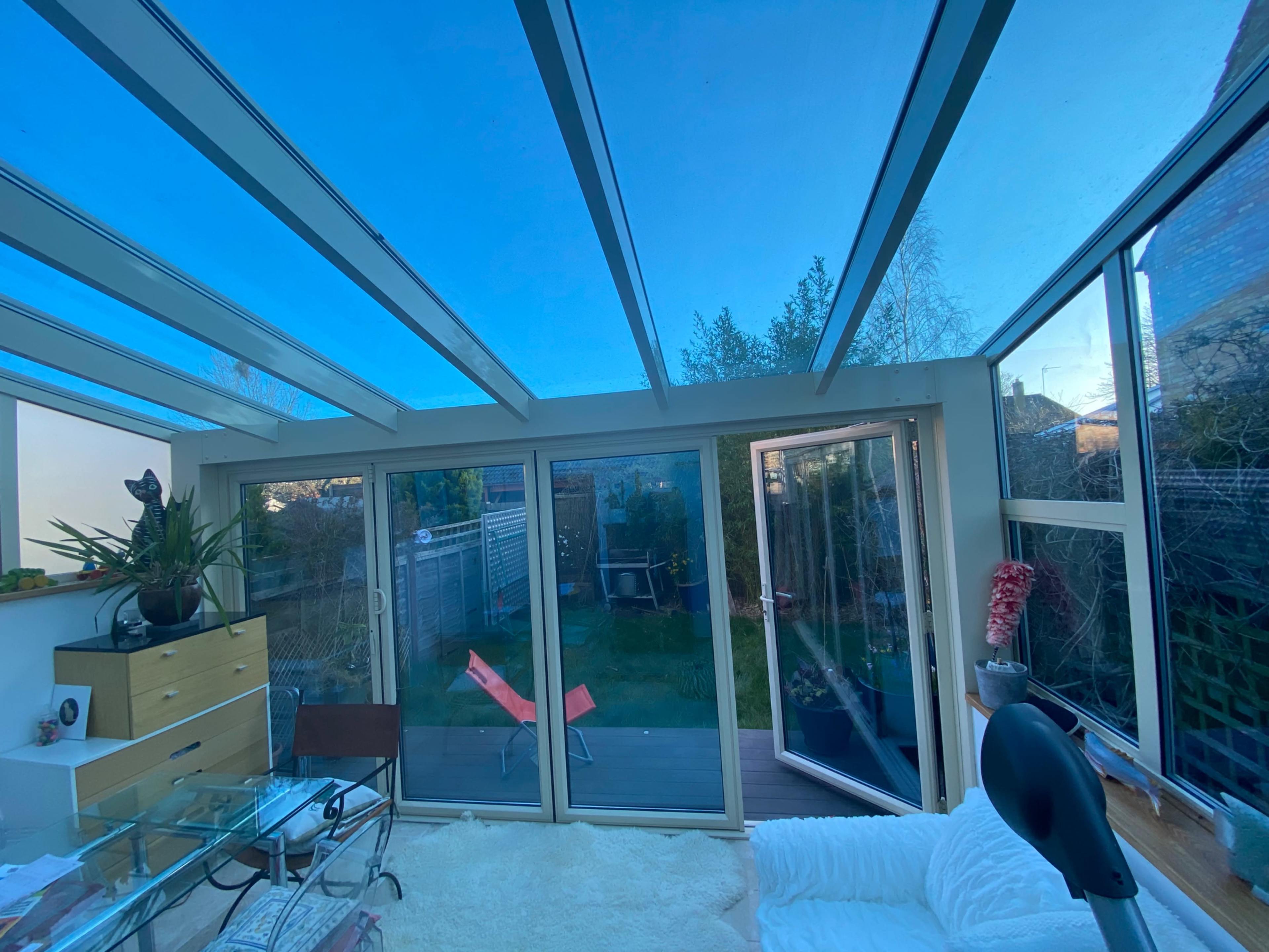 A sunroom with large glass walls and a ceiling, featuring modern furniture and a view of a garden outside.