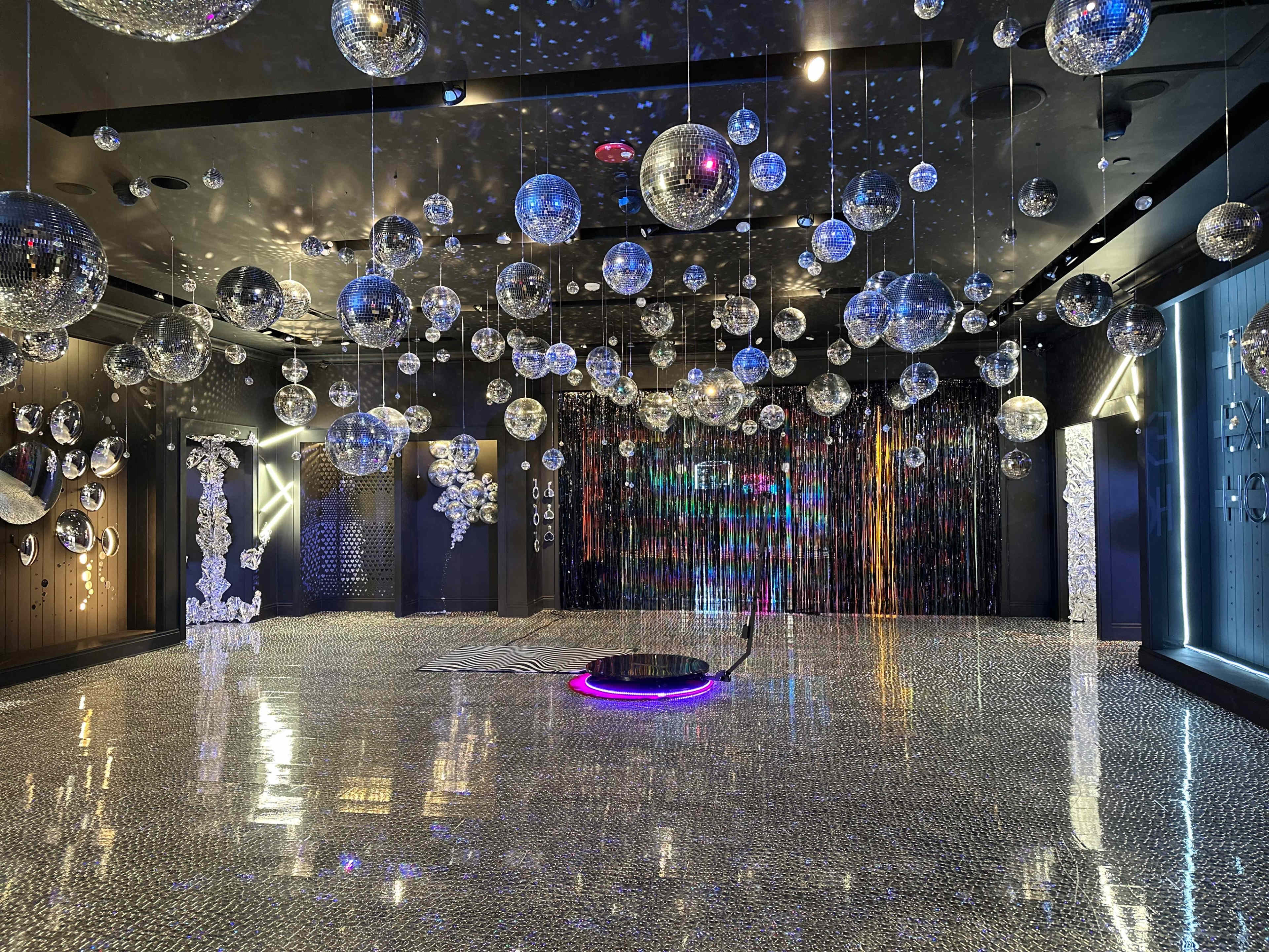 A spacious room adorned with numerous mirrored disco balls hanging from the ceiling, illuminated by colorful lights reflecting off a tiled floor.