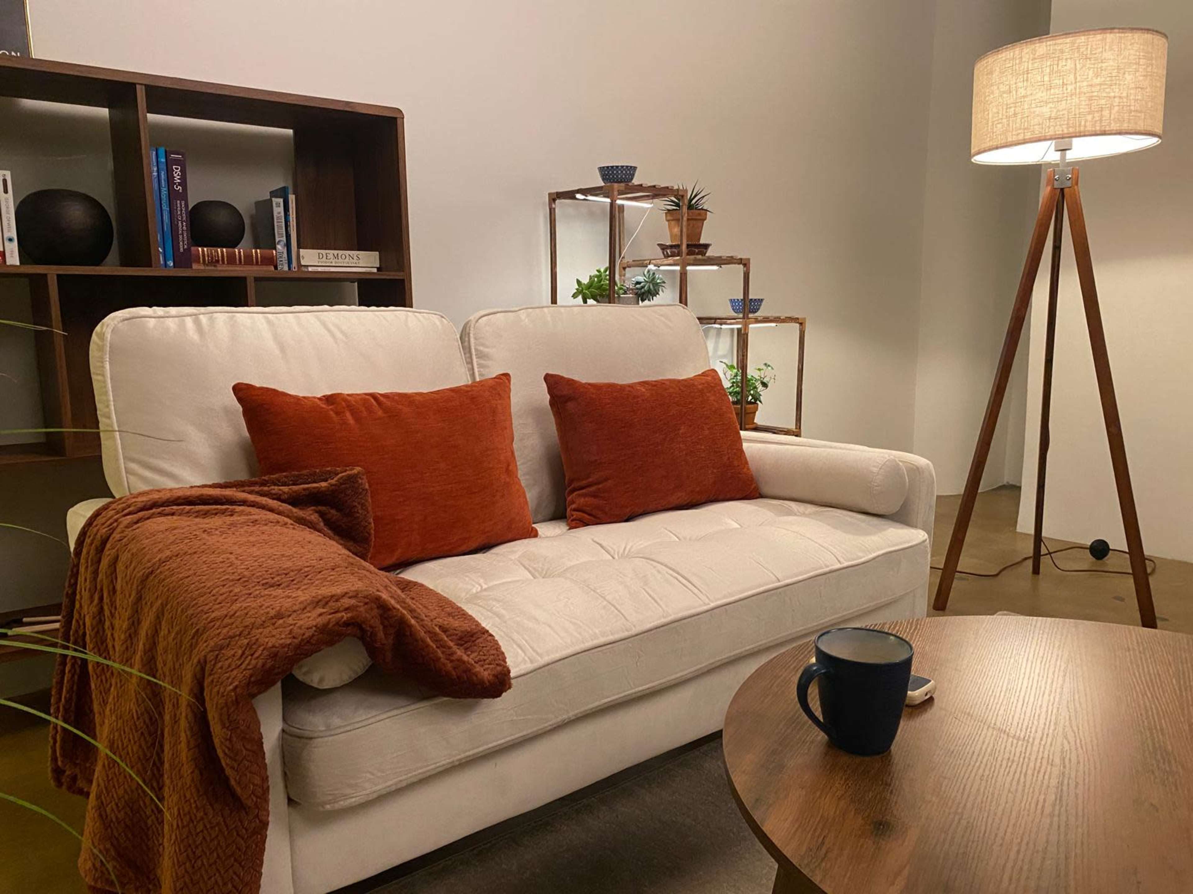 A white sofa with orange pillows is positioned next to a wooden side table and a floor lamp in a well-lit room adorned with plants and shelves.