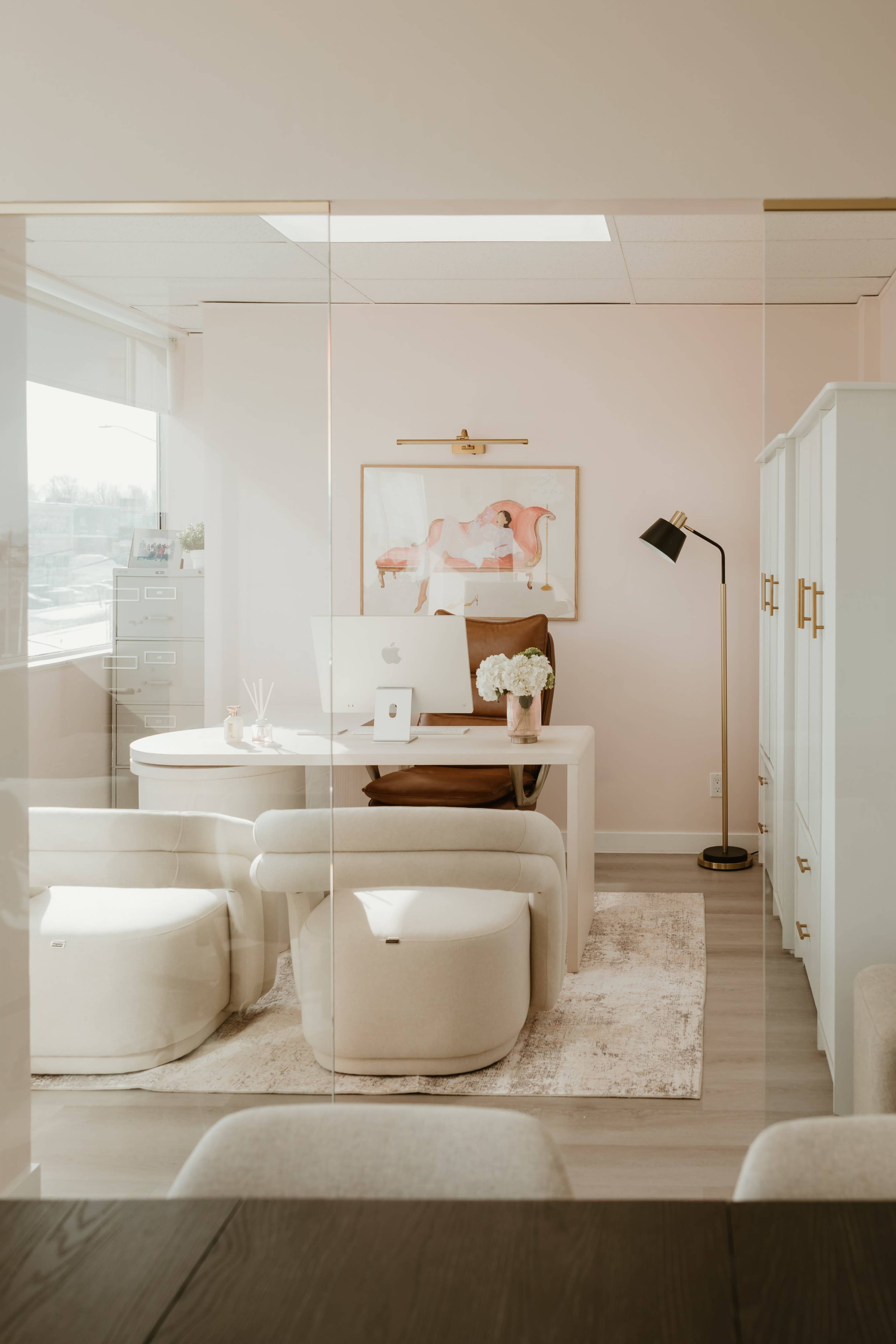 The image shows a modern office space with a white desk, a computer, and a framed artwork, all within a glass-enclosed area.