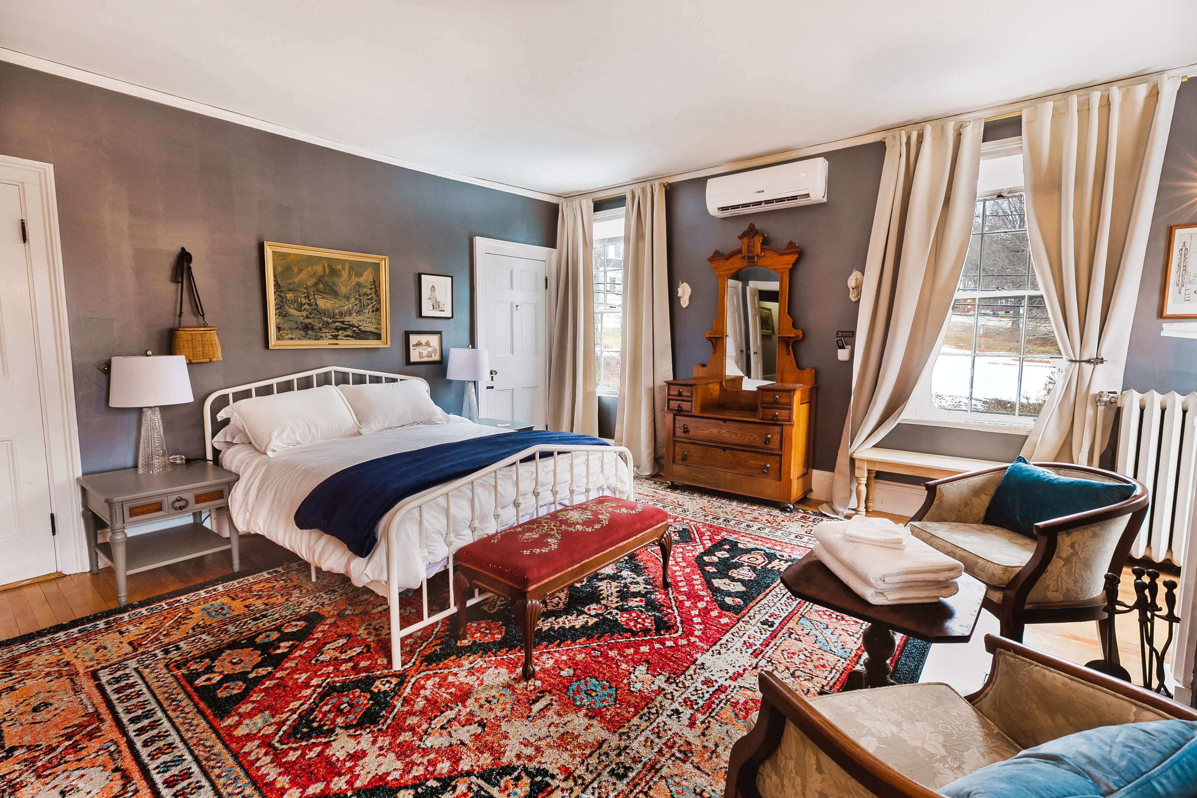 The image shows a cozy bedroom featuring a white bed, a wooden dresser, a vintage armchair, and a patterned rug on the hardwood floor, with natural light streaming in from large windows.