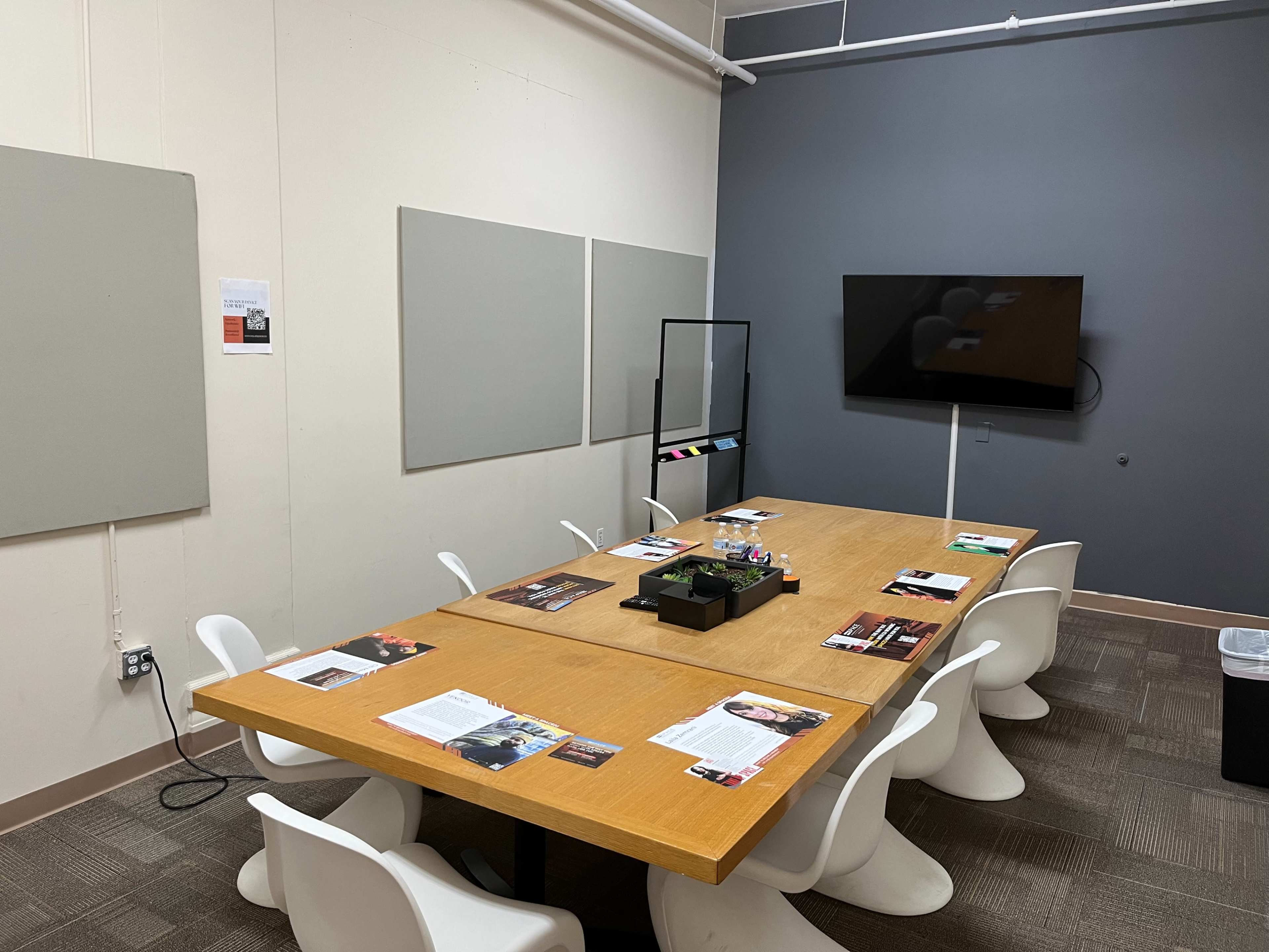 A meeting room is equipped with a large wooden table surrounded by white chairs, with wall-mounted screens and display materials on the table.