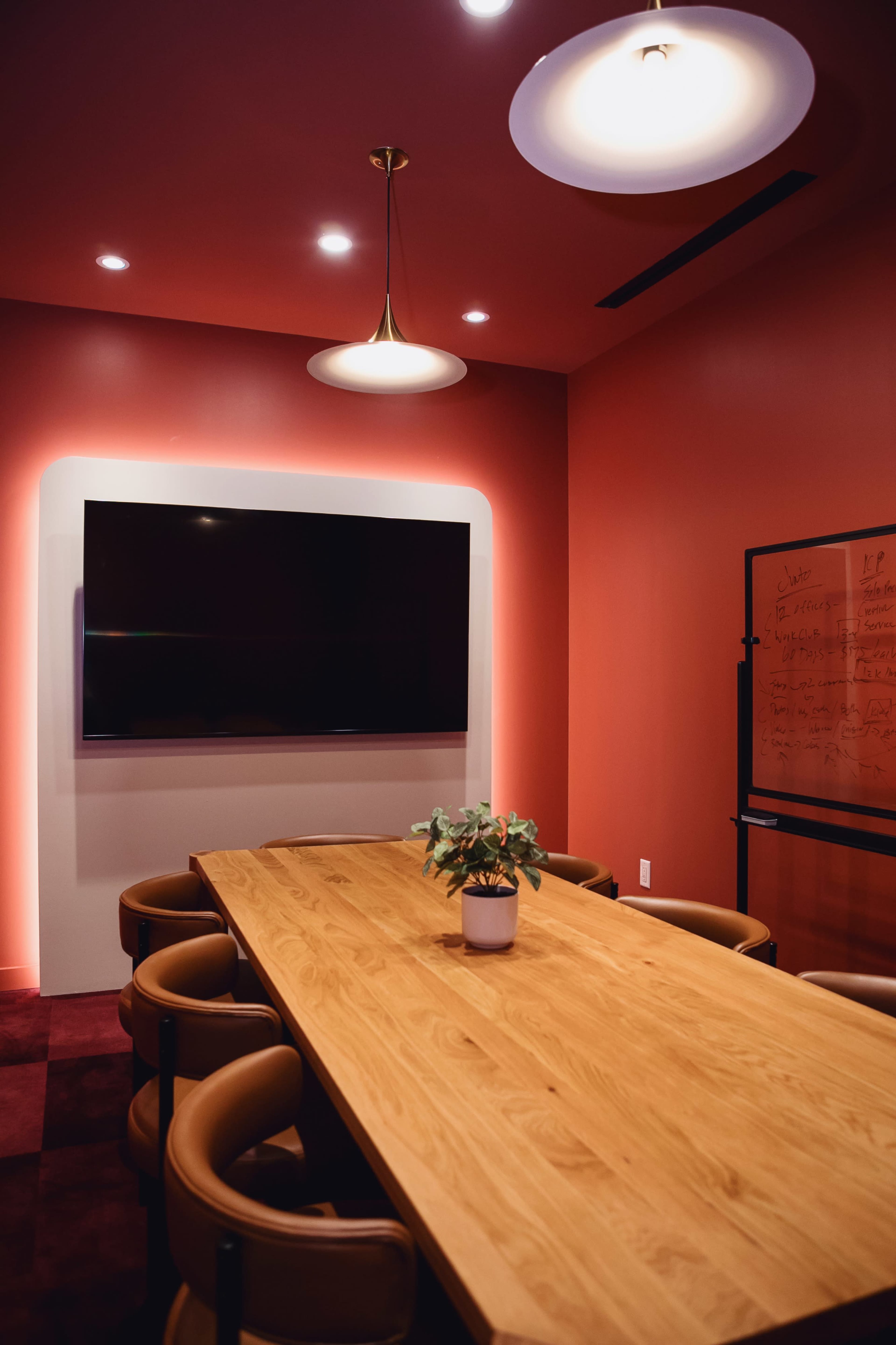 A modern conference room features a large wooden table surrounded by brown chairs, a wall-mounted television, and a whiteboard, all set against a red-painted wall.