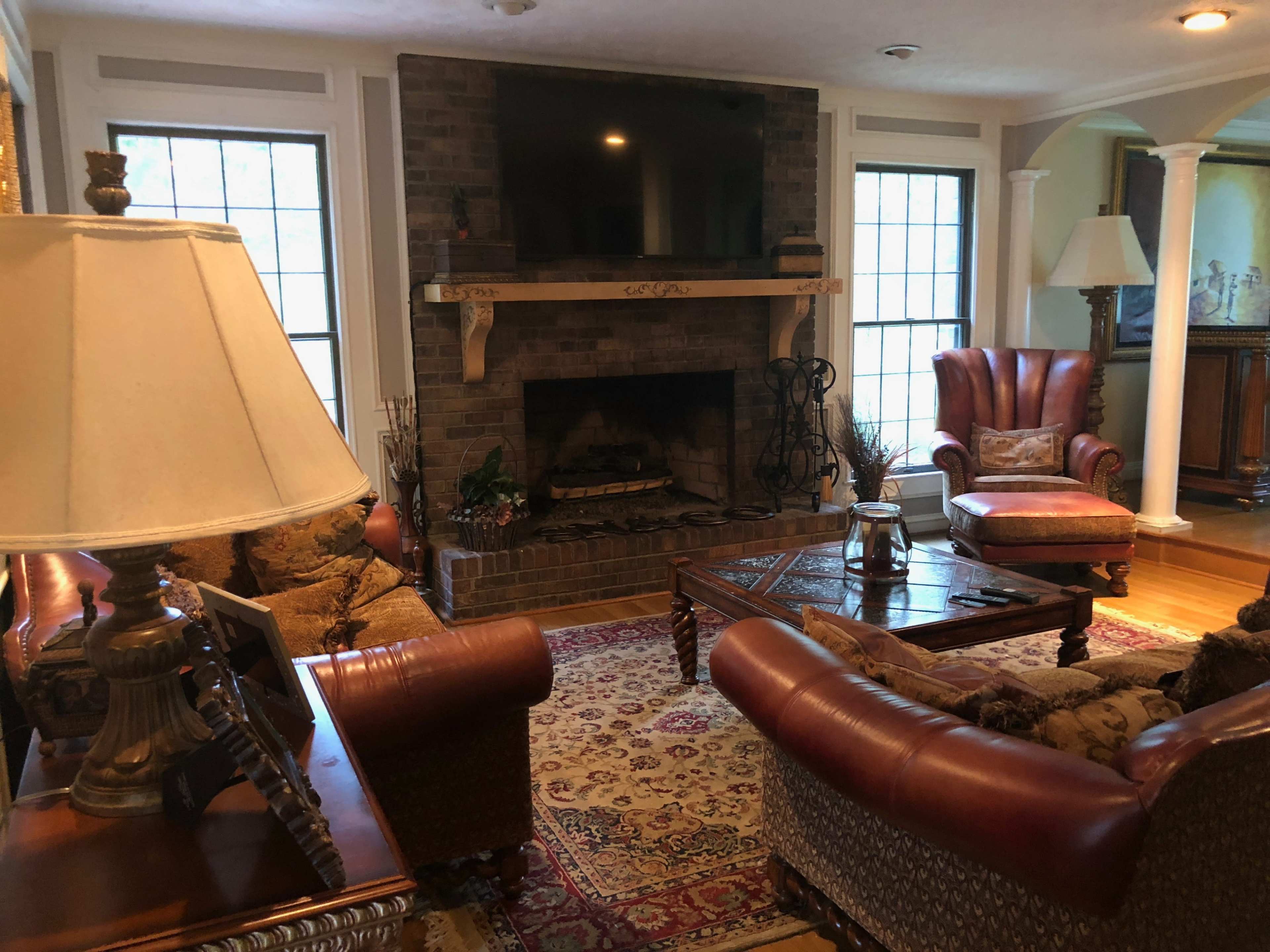 A furnished living room with a stone fireplace, two armchairs, and a coffee table on a patterned rug, illuminated by natural light from large windows.