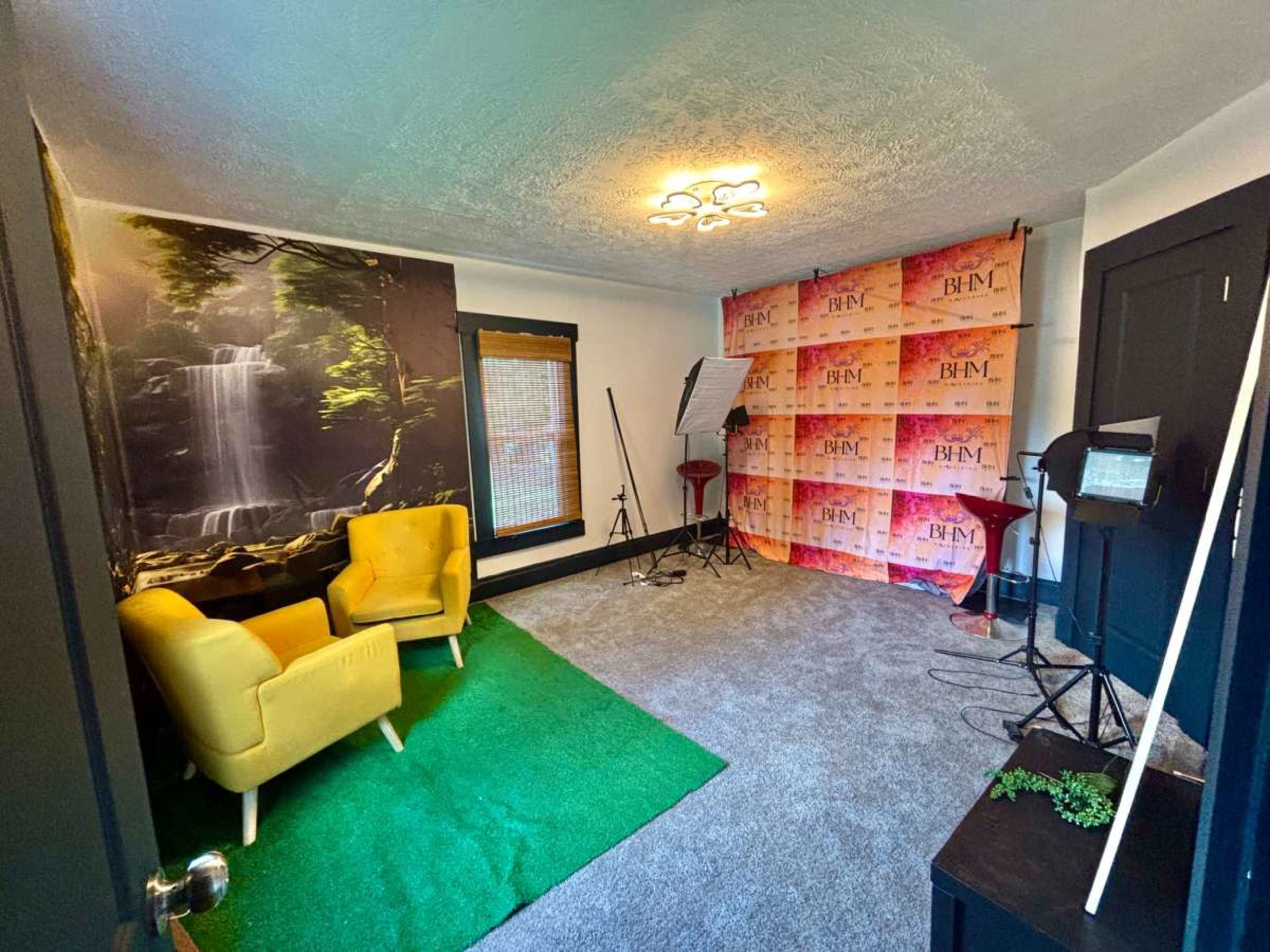 The image shows a small studio space with yellow chairs, a green carpet, a backdrop with a brand logo, and lighting equipment set up for photography or video production.