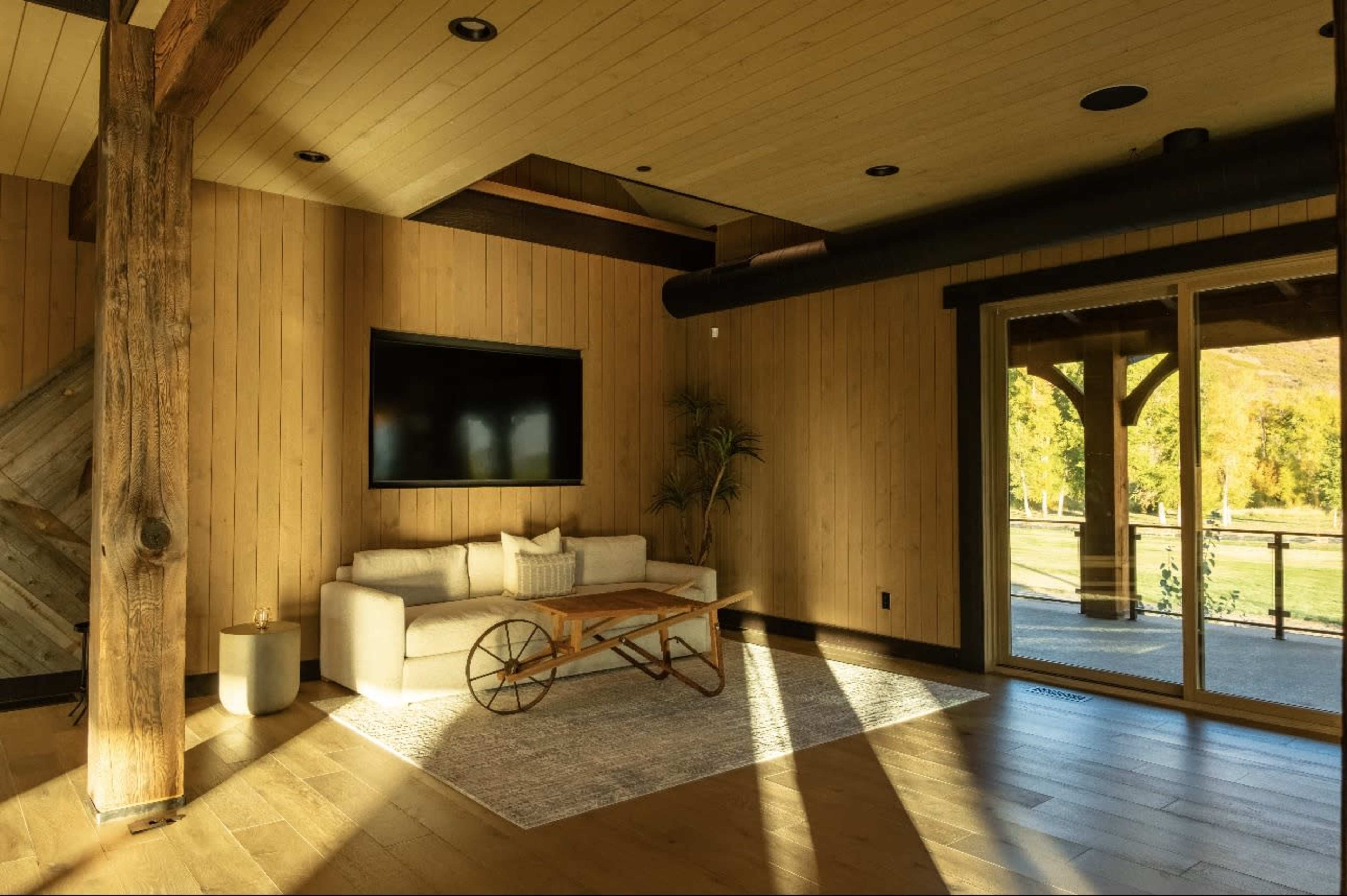 The image shows a cozy living room with wooden walls, a television mounted on the wall, a white sofa, a coffee table, and large windows that provide a view of the outdoors.