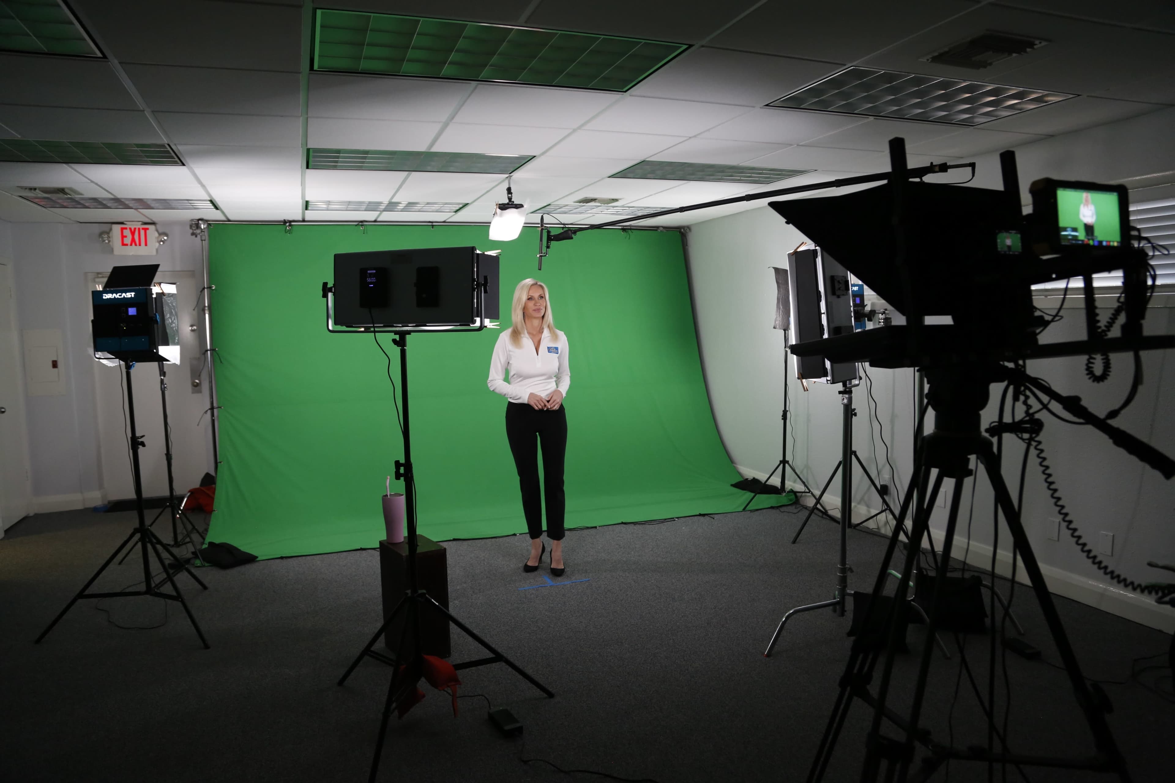 A woman stands in front of a green screen in a studio, surrounded by cameras and lighting equipment.