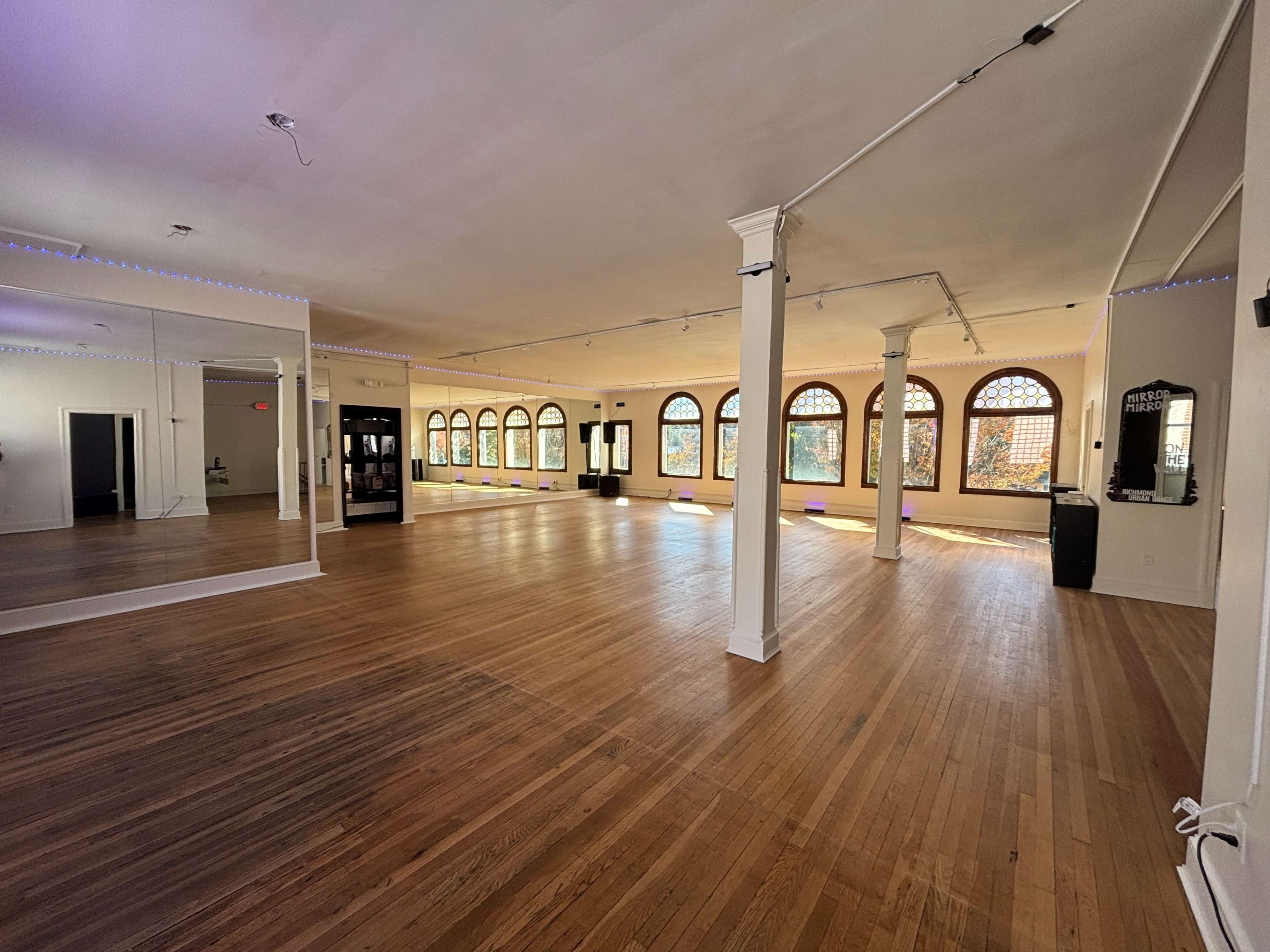 The image shows a spacious, empty room with wooden flooring and large arched windows along one wall.