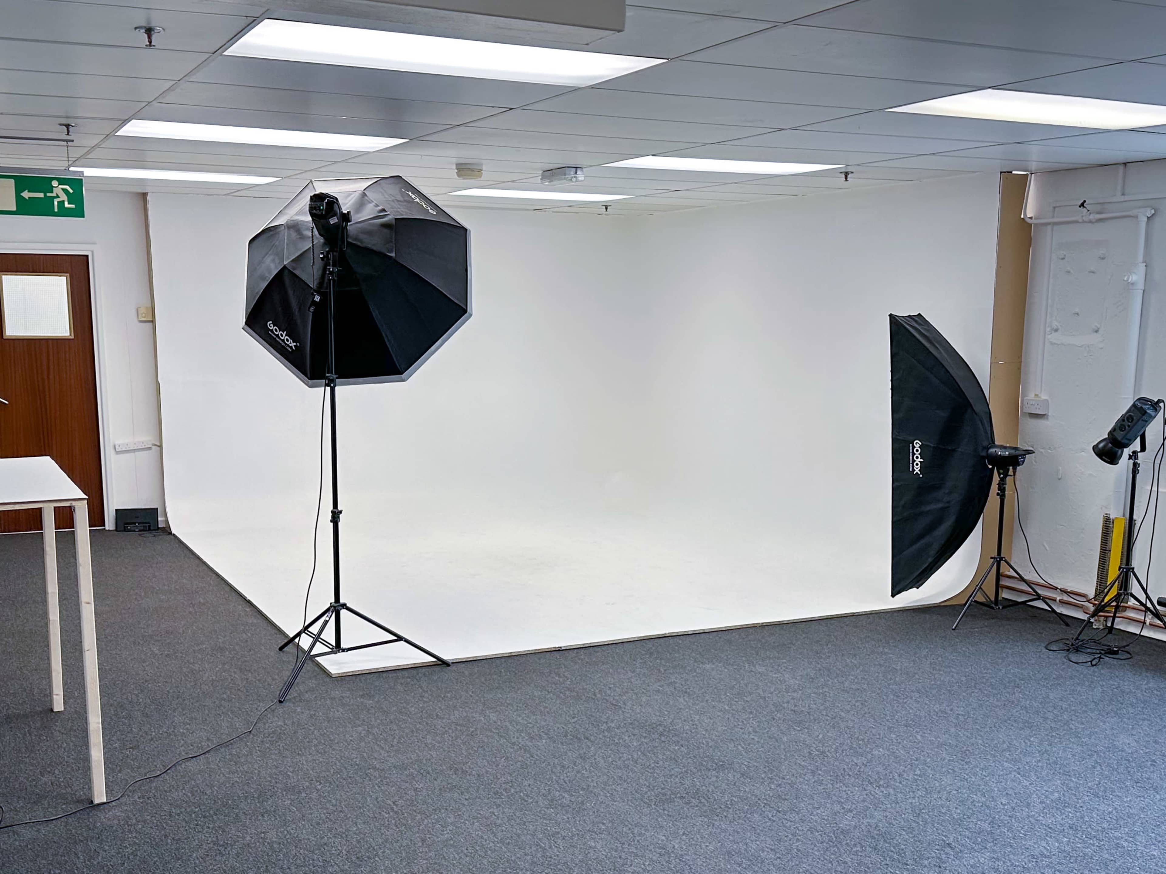 A photography studio with a white backdrop, two softbox lights on tripods, and a gray carpeted floor.
