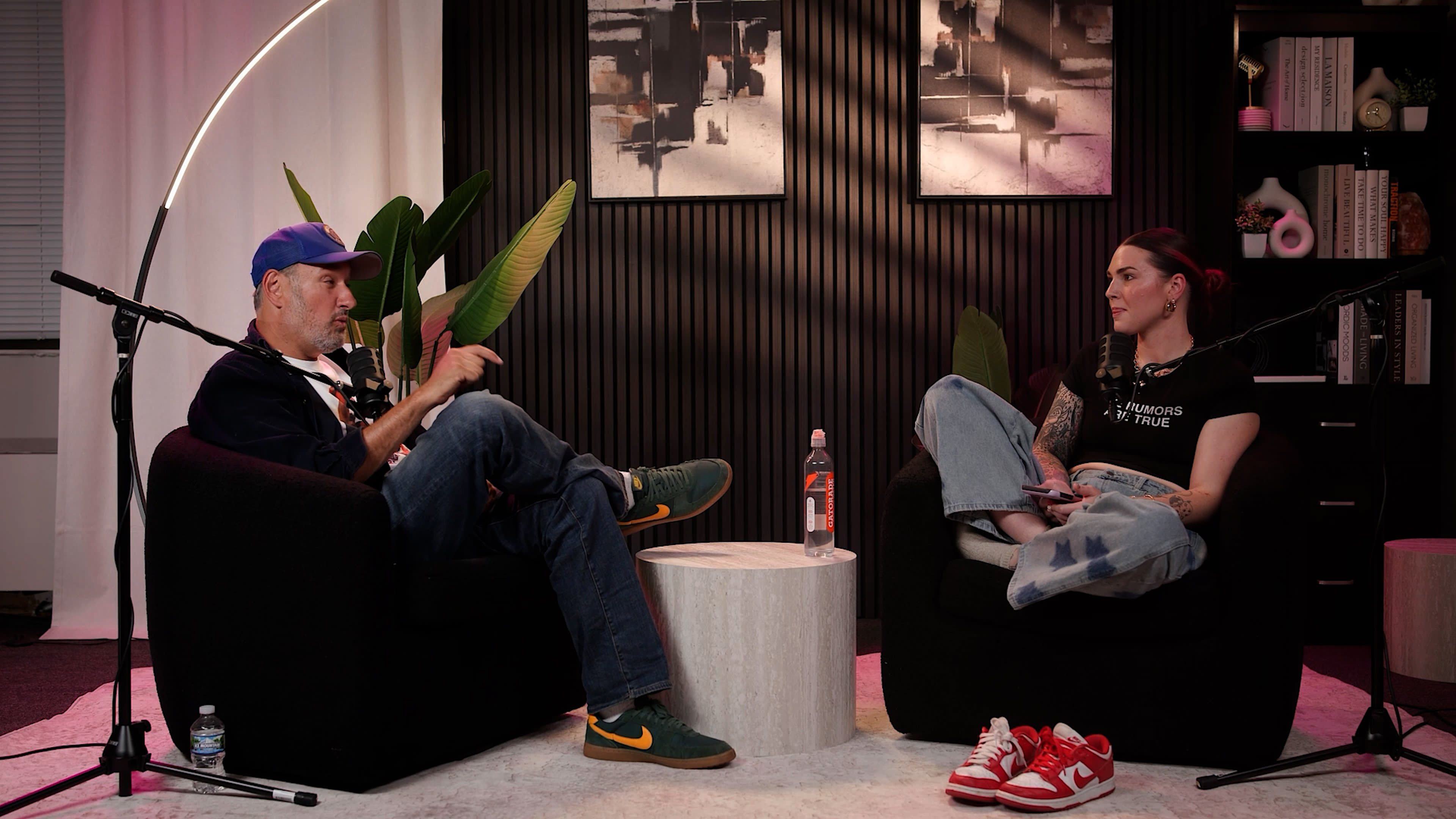 A man and a woman are seated in modern armchairs, engaged in conversation, surrounded by indoor plants and abstract artwork on the wall.