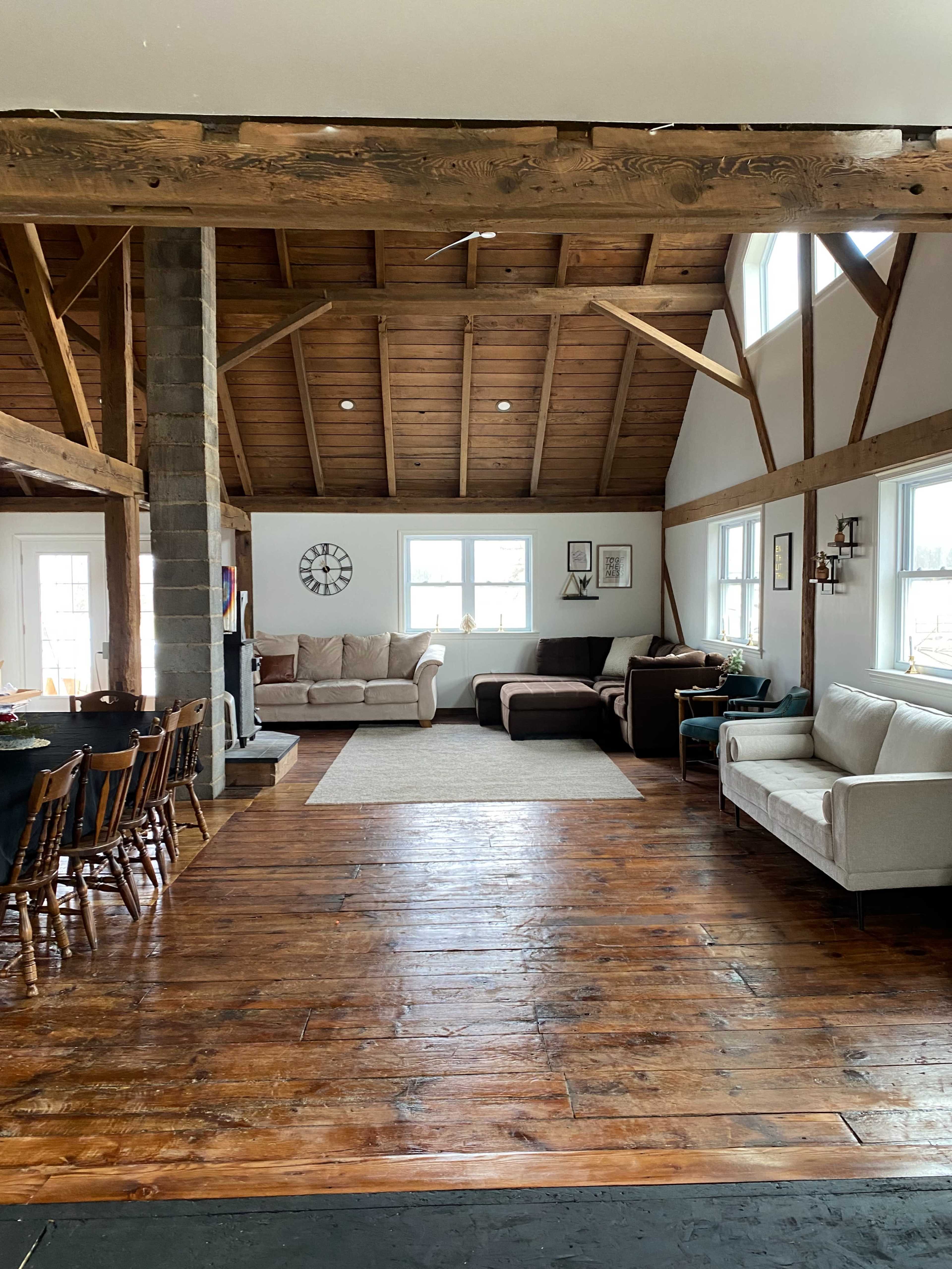 A spacious, well-lit living area with wooden beams, a rustic wood floor, multiple sofas, and a dining table with chairs.