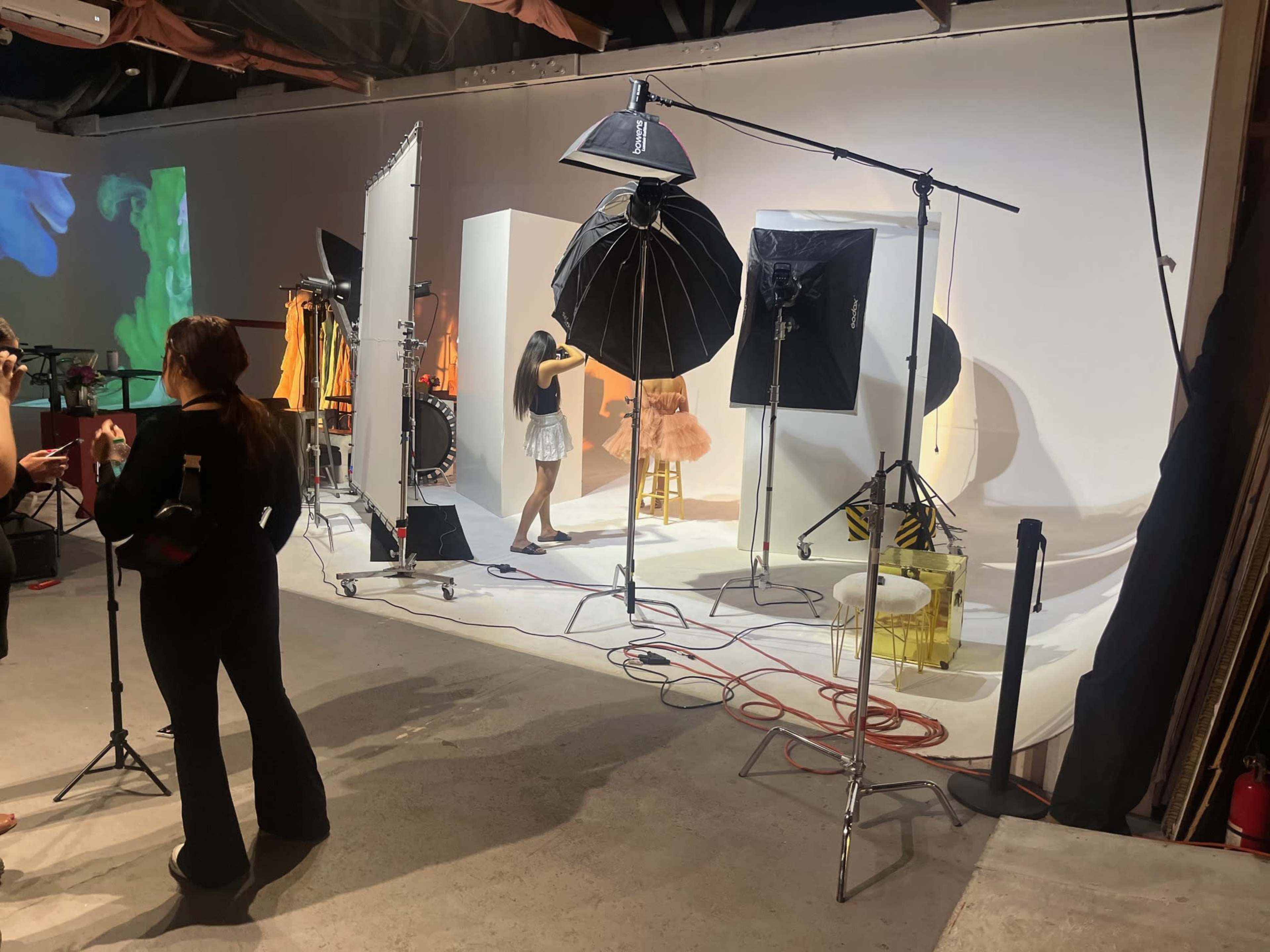 The image shows a photography studio set up with various lighting equipment, backdrops, and a model posing on a stool.