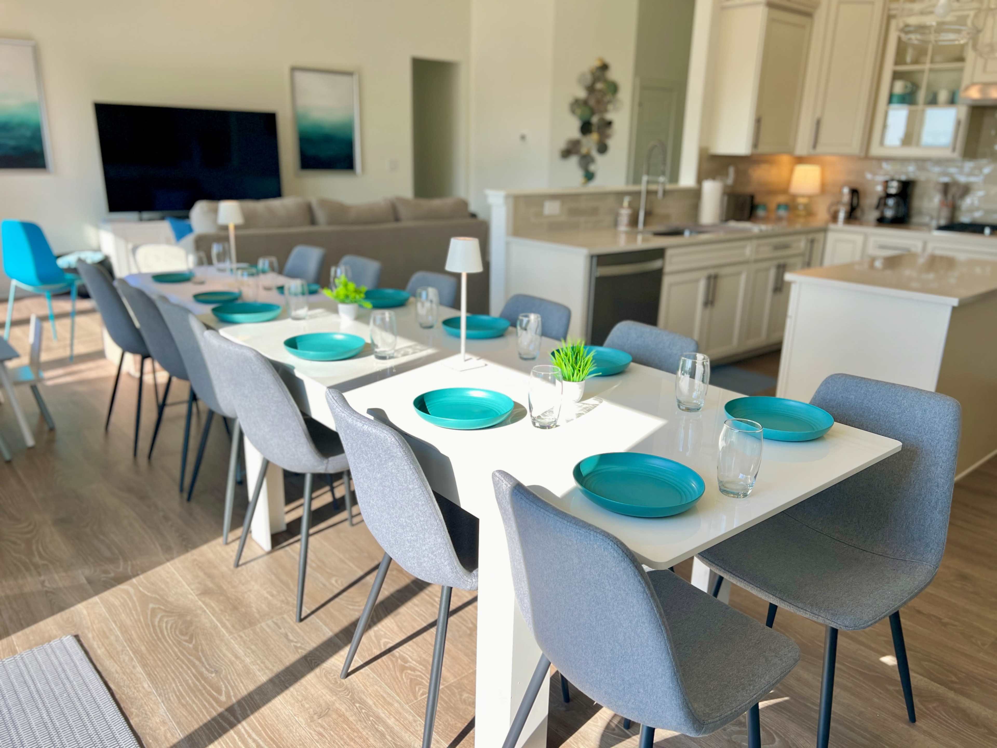 A modern dining area features a long table set with turquoise plates and glasses, surrounded by gray chairs, with a kitchen and living space visible in the background.