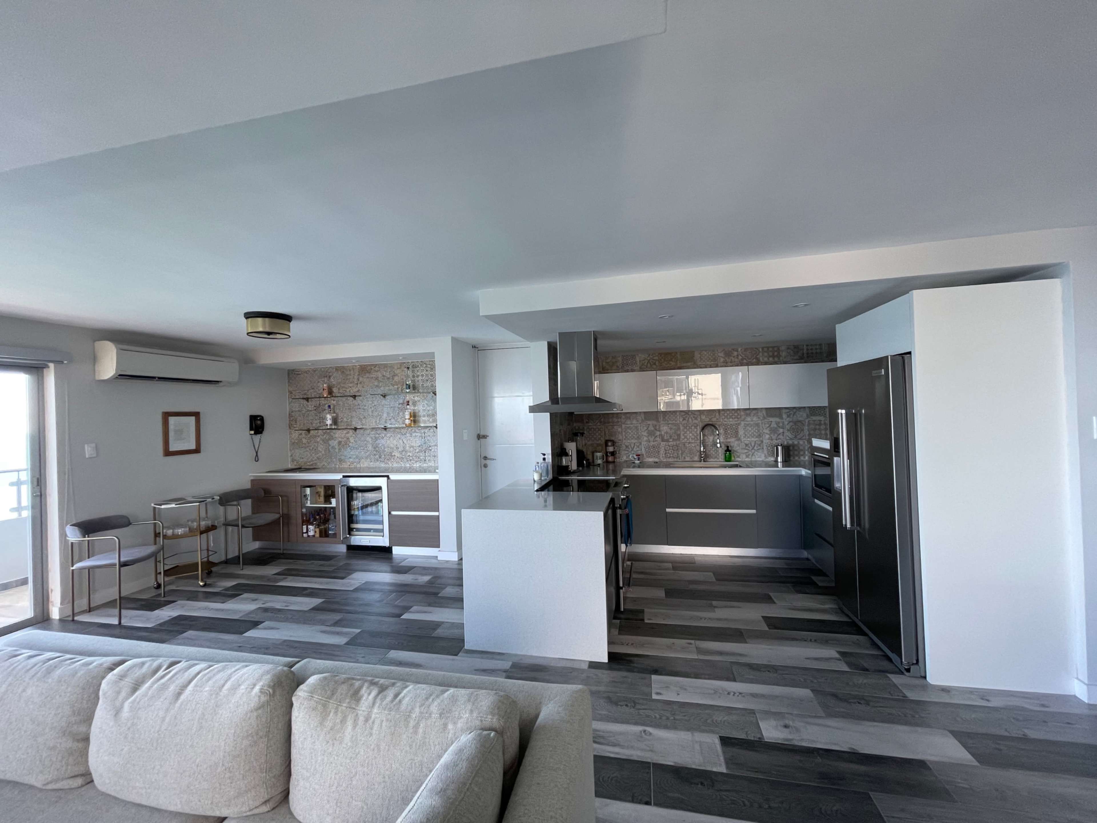 The image shows a modern kitchen and living area with a light-colored sofa, sleek cabinetry, and a dining bar.